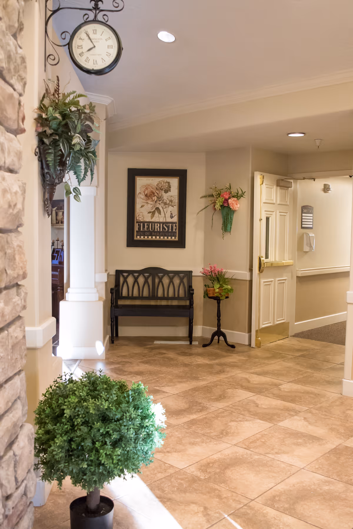 A well-lit interior hallway area with beige tiled flooring and cream-colored walls. There is a black bench against the wall beneath a framed floral artwork labeled 'Fleuriste'. To the right of the bench is a small table with a flower arrangement and a wall-mounted green vase with flowers. A round clock with Roman numerals hangs from a decorative bracket on the left wall near a stone pillar. A potted green plant is in the foreground on the left side. An open door leads to a hallway with a directory sign on the wall.