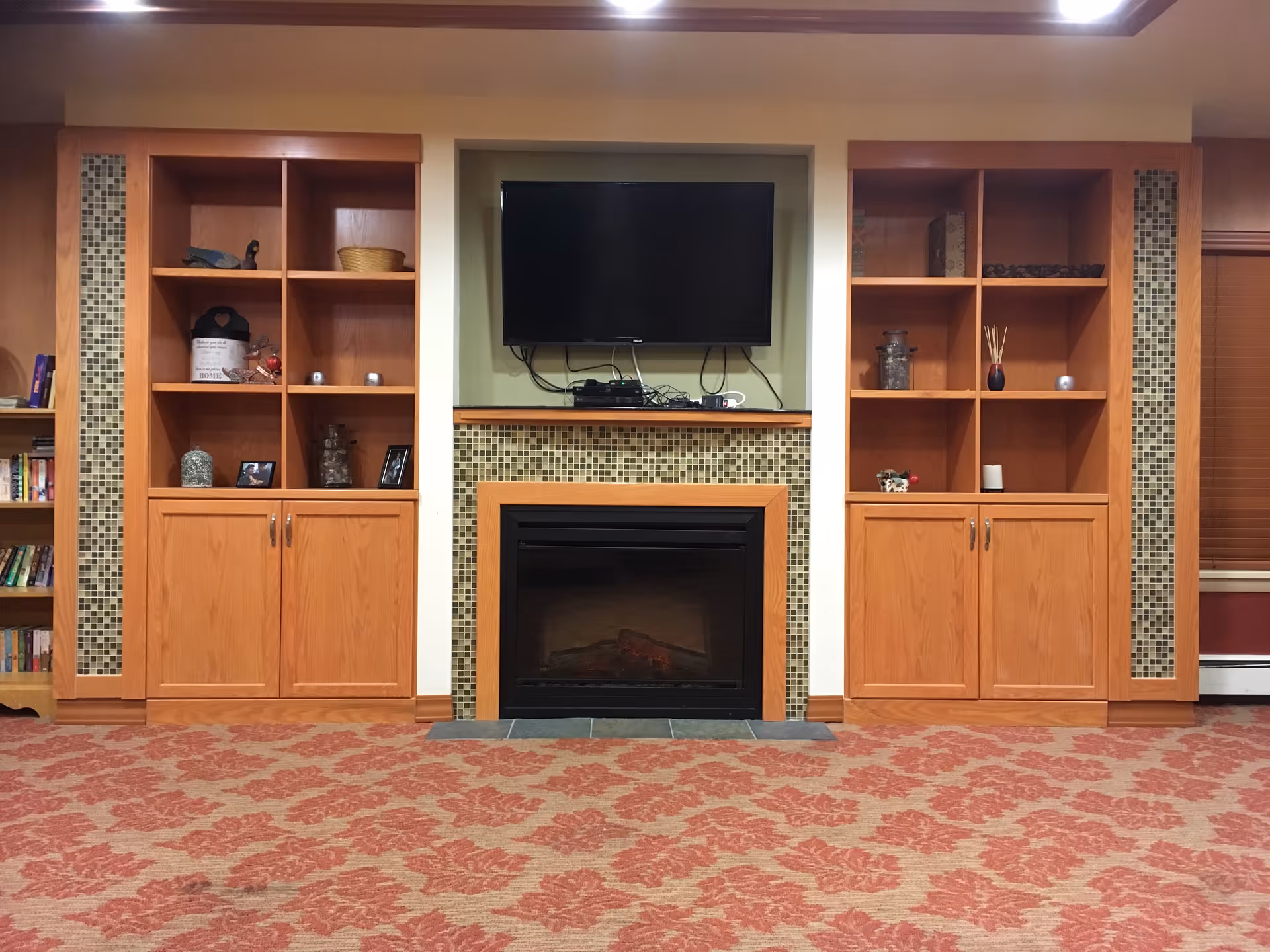 A cozy living room area featuring a fireplace with a mosaic tile surround and a mounted flat-screen TV above it. On either side of the fireplace are wooden built-in shelves and cabinets with various decorative items and books. The floor is carpeted with a red and beige floral pattern.