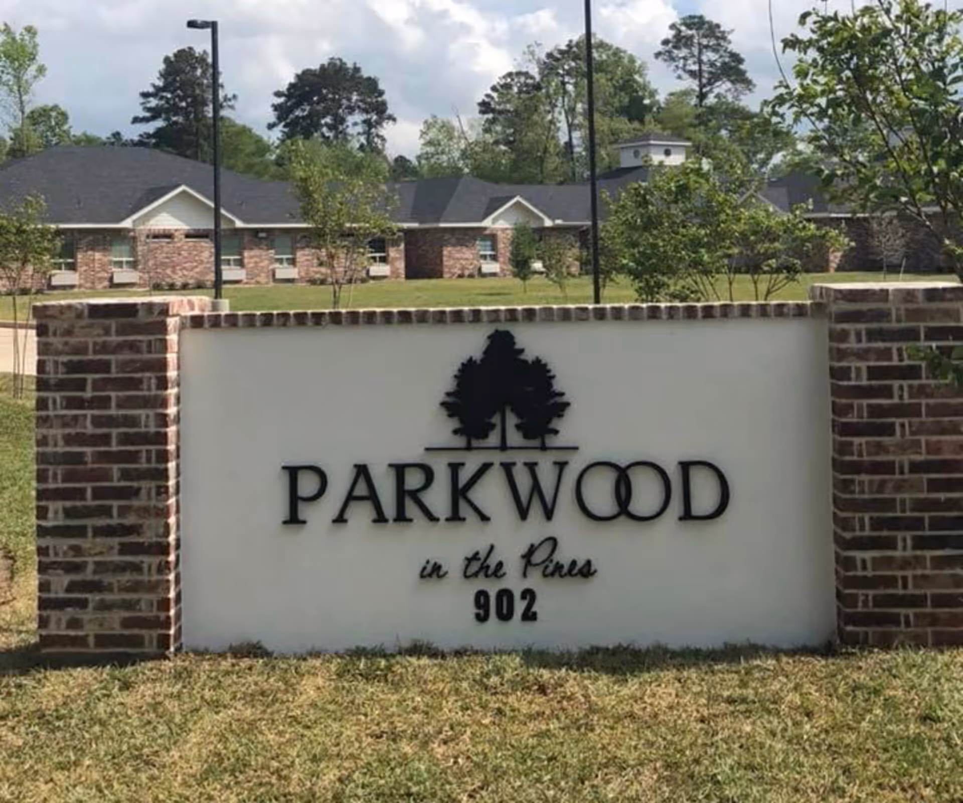 Entrance sign reading "PARKWOOD in the Pines 902" with a tree logo in front of a brick building and lawn.