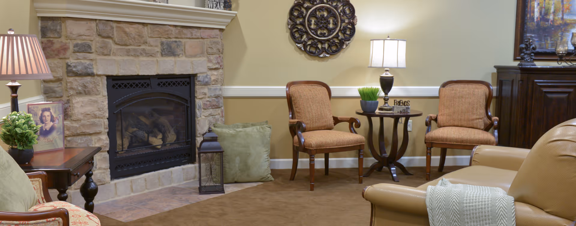 A cozy living room area with a stone fireplace, two upholstered chairs, a small round table with a lamp and plant, a leather sofa with a throw blanket, and decorative wall art above the fireplace and table.