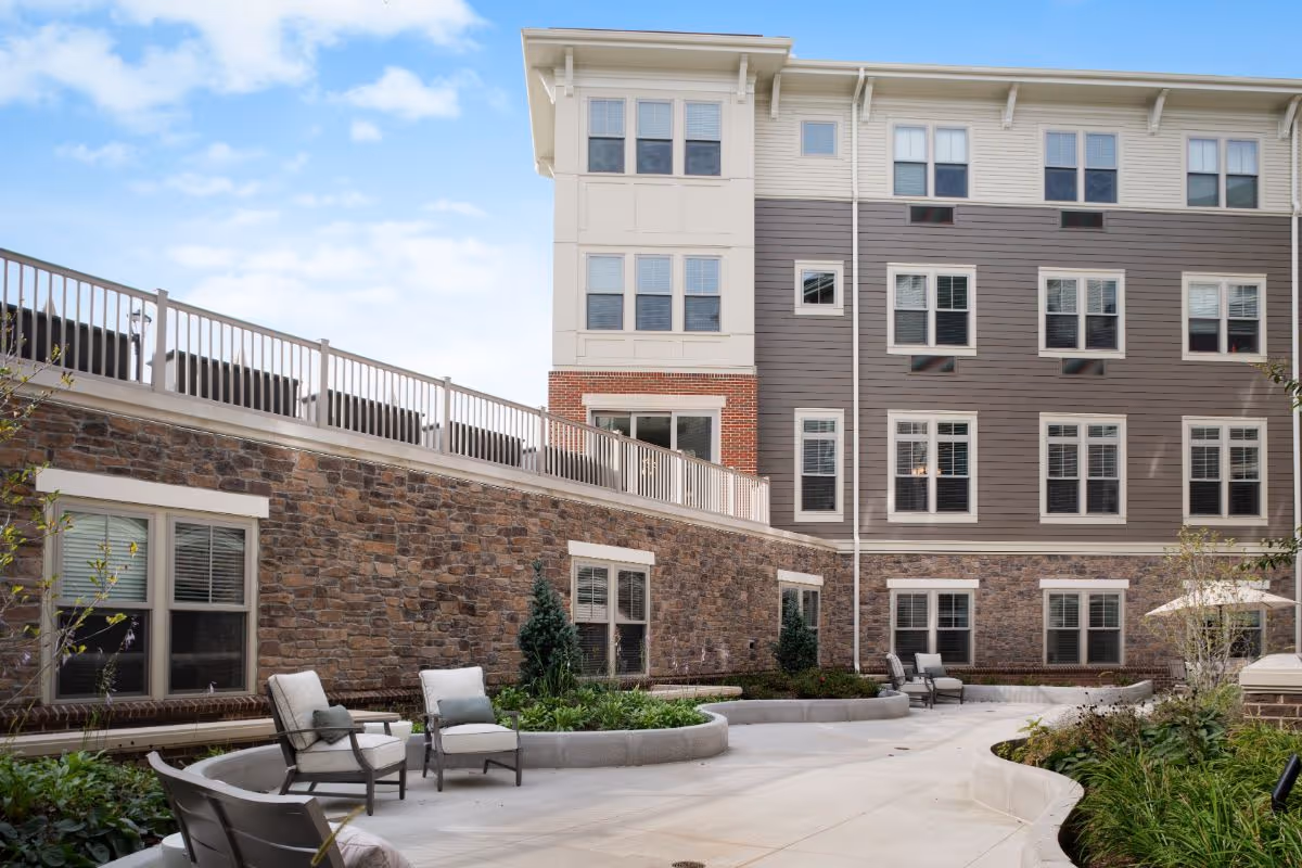 Outdoor courtyard of a multi-story senior living building with patio seating, planters, and a stone-and-siding exterior.