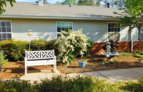 Outdoor garden area at Gaines Park Senior Living featuring a white wooden bench, a flowering bush, a potted plant, a birdbath with a small statue, and a paved walkway in front of a beige building with windows.