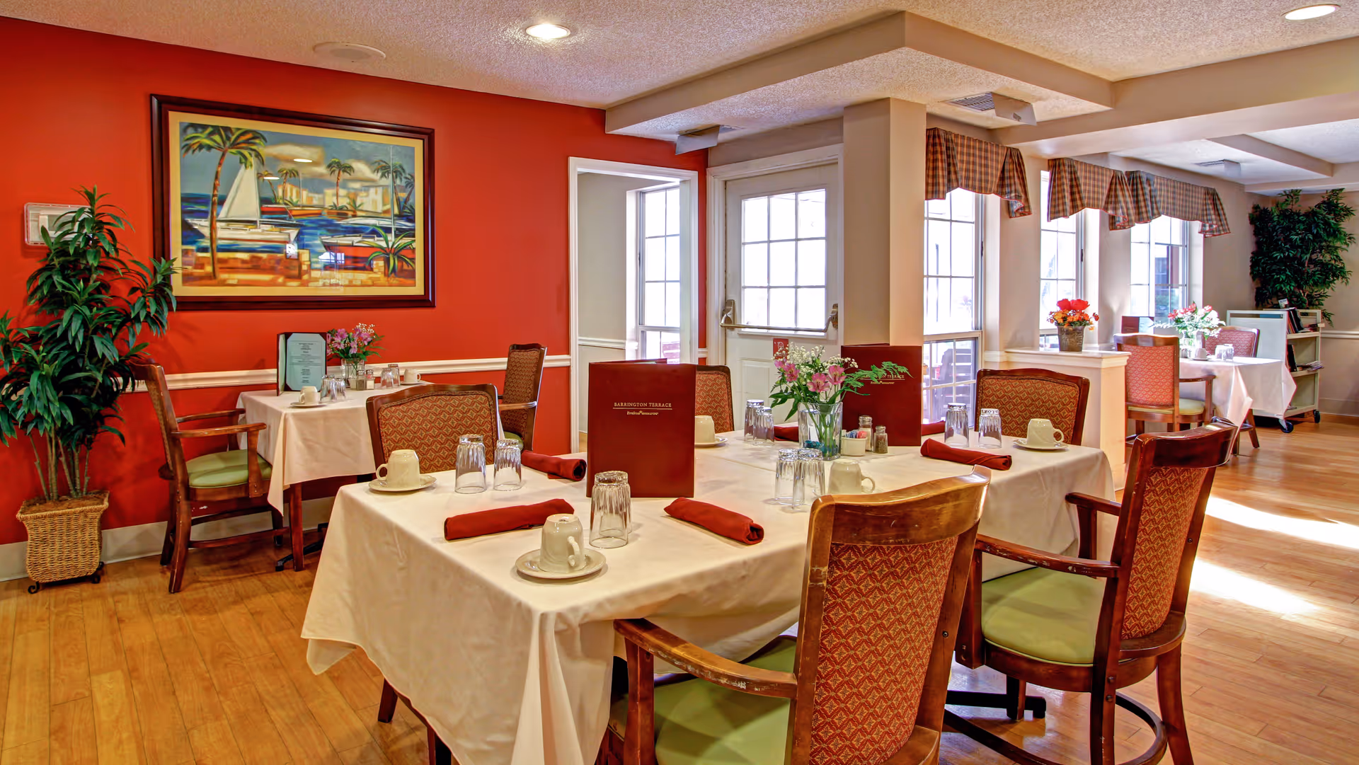 Well-lit dining room with tables set for meals, upholstered chairs, table linens, and floral centerpieces.
