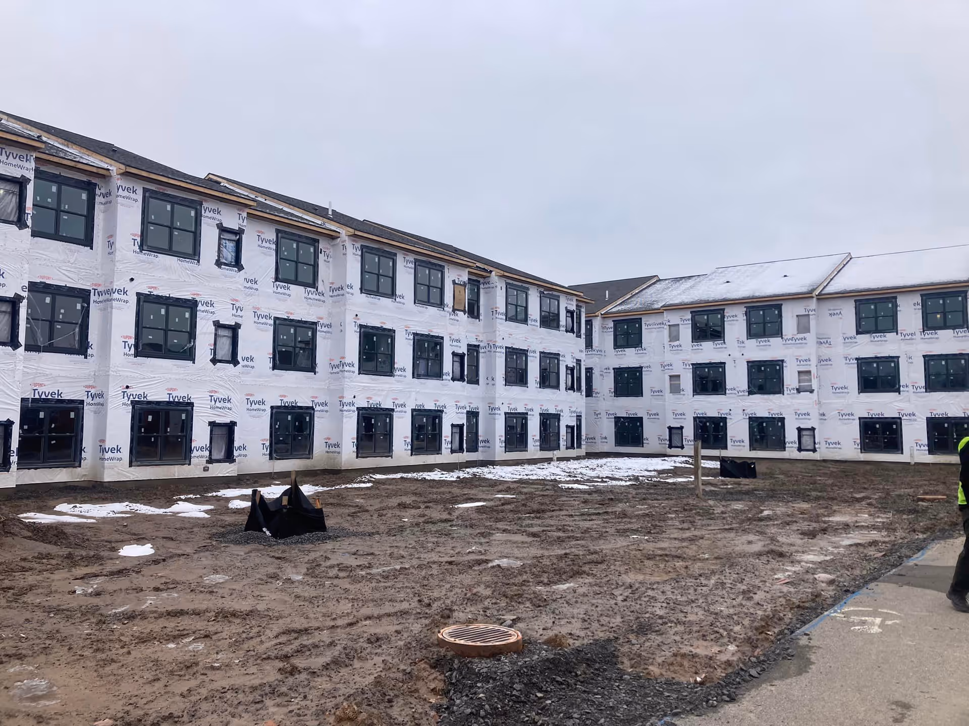 Exterior view of a three-story senior living building under construction wrapped in Tyvek with a muddy foreground and patches of snow.