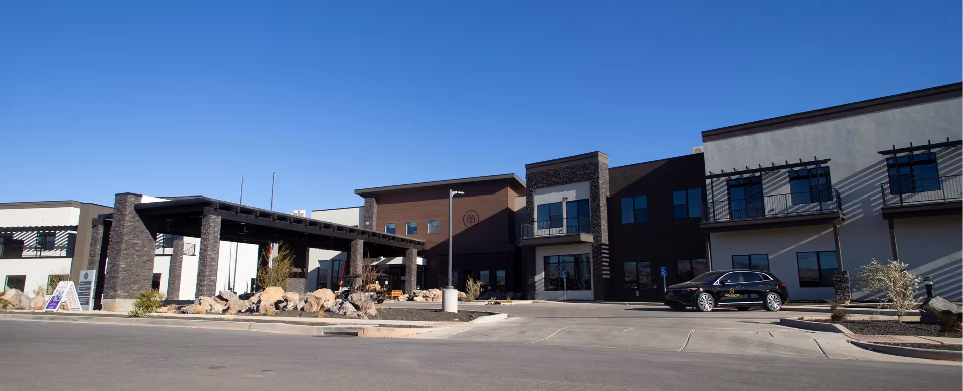 Exterior view of Snow Canyon Retirement Community building under a clear blue sky. The building features modern architecture with stone pillars, large windows, balconies, and a covered entrance. A black vehicle with the community's logo is parked near the entrance.