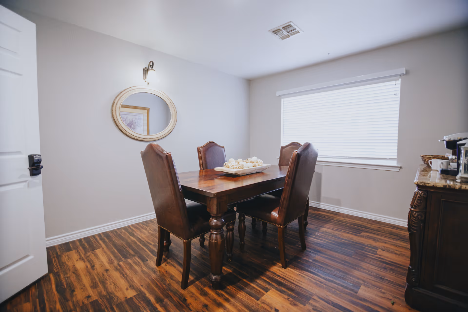 Wooden dining table with four leather chairs in a bright room with hardwood floors and a round wall mirror.