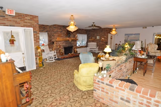 A cozy living room area in an assisted living facility featuring patterned carpet, brick walls, a brick fireplace decorated with garlands, rocking chairs, upholstered armchairs, and various lamps and floral decorations. The room has a warm and inviting atmosphere with ceiling fans and an exit door visible on the left.