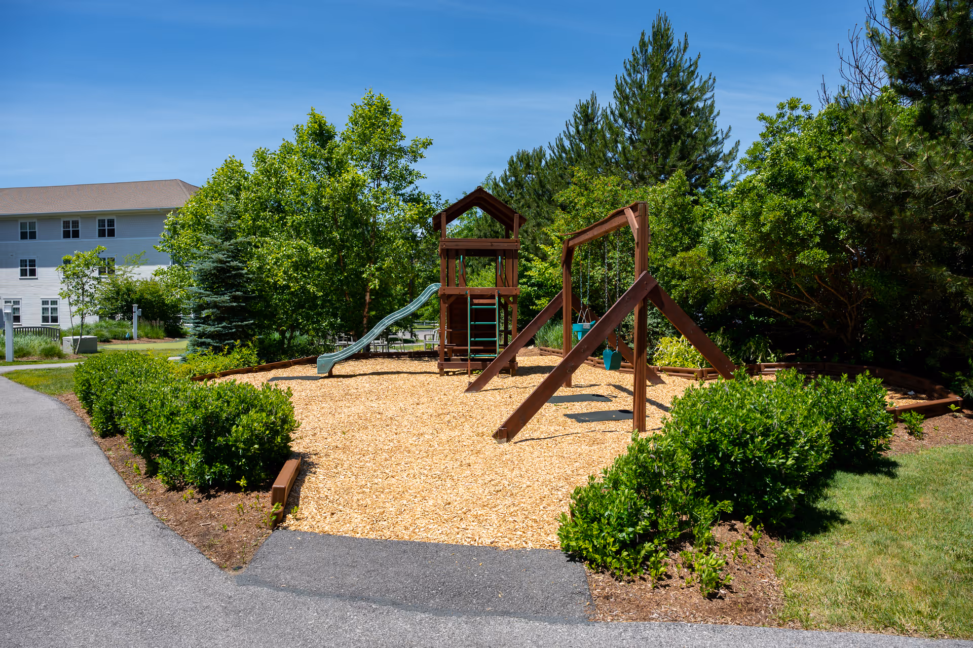 Outdoor playground area with a wooden play structure including a slide and swings, surrounded by wood chips and greenery. A paved pathway and bushes border the playground, with trees and a multi-story building visible in the background under a clear blue sky.