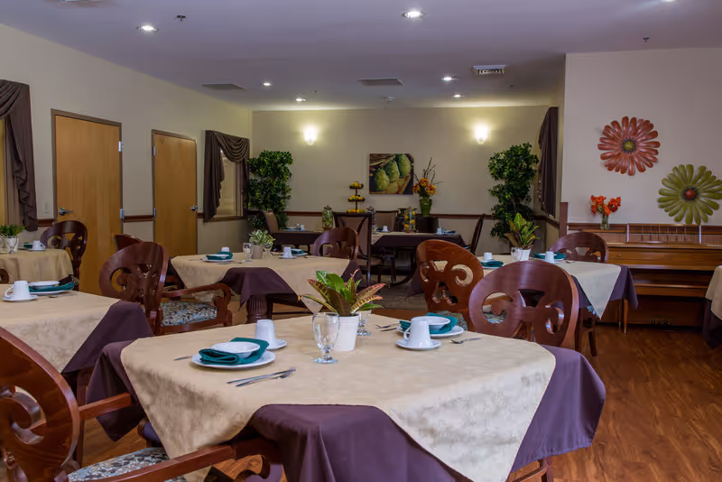 A dining room in a senior living facility with multiple tables covered in beige and purple tablecloths, each set with plates, cups, utensils, and napkins. Wooden chairs surround the tables, and there are plants and floral decorations on the walls and tables. The room has wooden flooring and soft lighting.