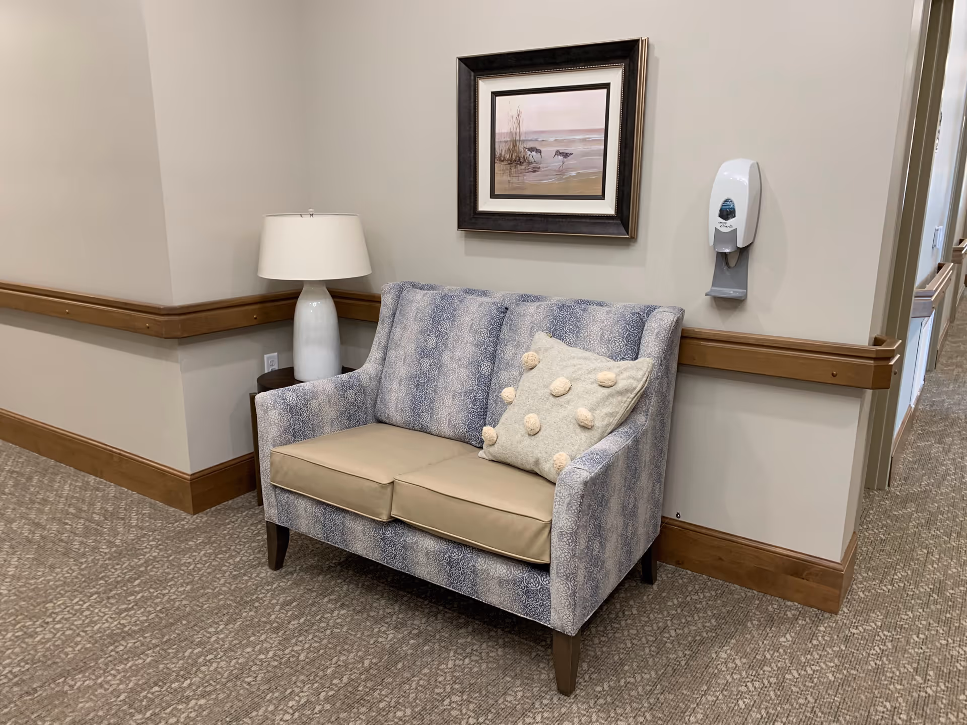 A small seating area in a hallway with a patterned loveseat featuring beige cushions and a decorative pillow with pom-poms. Next to the loveseat is a white table lamp on a small side table. Above the loveseat hangs a framed picture of birds near water. A wall-mounted hand sanitizer dispenser is visible on the right wall. The walls are light-colored with wooden chair rails and baseboards, and the floor is carpeted.