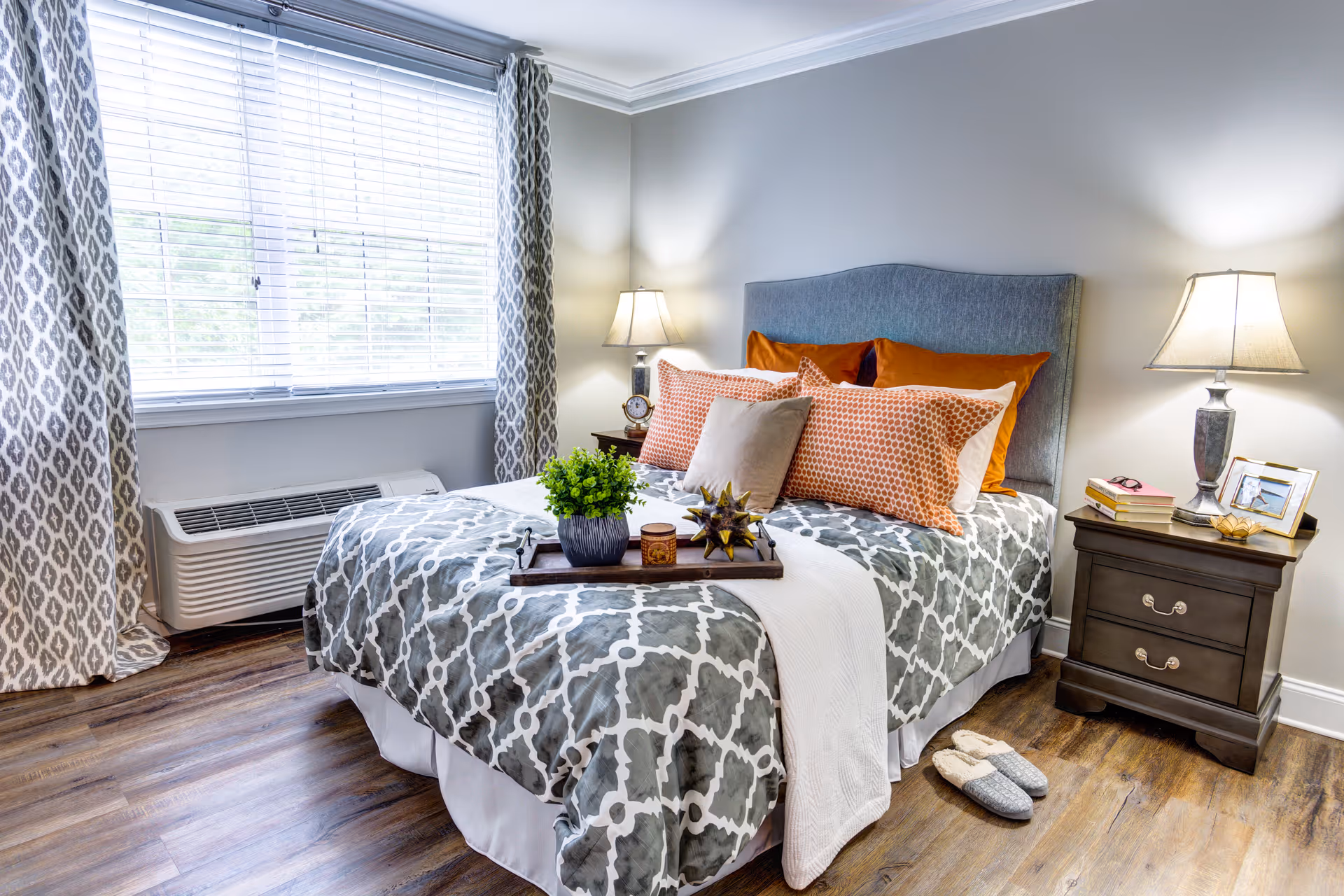 A cozy bedroom with a neatly made bed featuring a gray and white patterned comforter, orange and beige pillows, and a gray upholstered headboard. There are two bedside tables with lamps, books, and decorative items. A window with patterned curtains lets in natural light, and a pair of slippers is placed on the wooden floor near the bed.