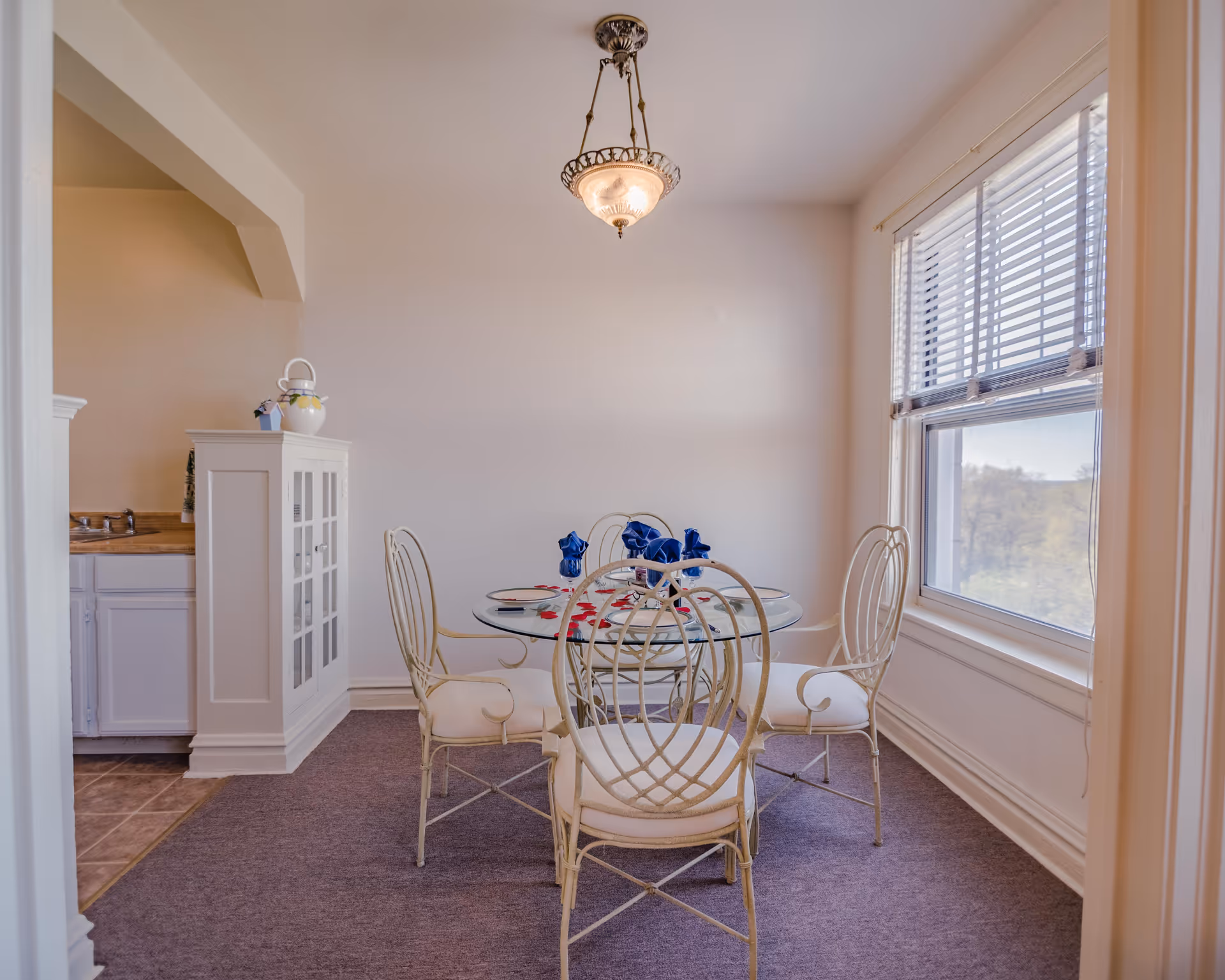 A bright dining area with a round glass table set for four with white cushioned metal chairs. The table is decorated with blue napkins and scattered red rose petals. A large window with blinds lets in natural light, and a vintage-style ceiling light fixture hangs above the table. To the left, part of a kitchen sink and a white cabinet with glass doors are visible.