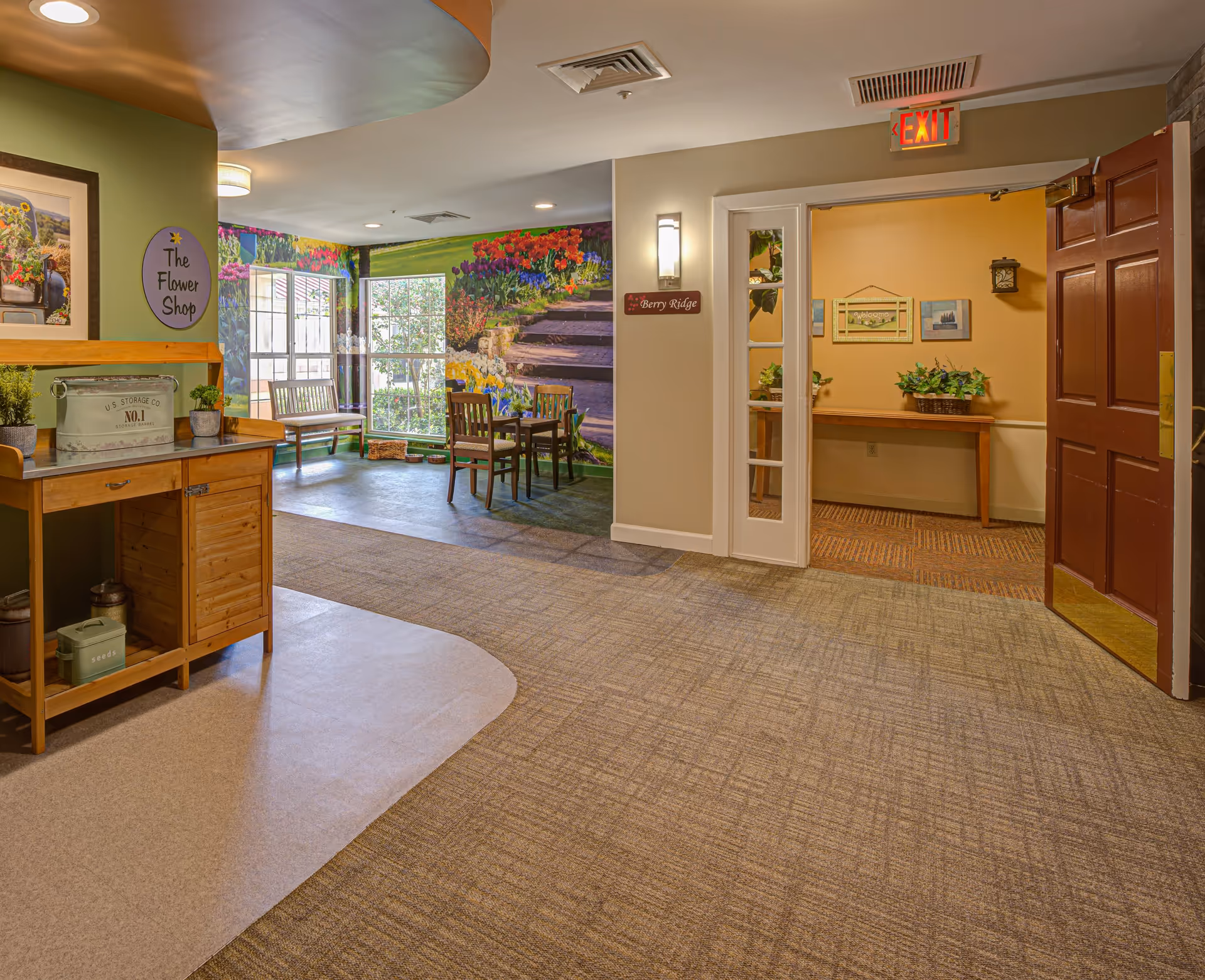 Interior view of a senior living facility hallway with a small seating area featuring a table and chairs near a window with a colorful floral mural. A wooden cabinet labeled 'The Flower Shop' is on the left side, and an open door on the right leads to another room with a welcome sign and plants on a wooden table. An exit sign is visible above the door.
