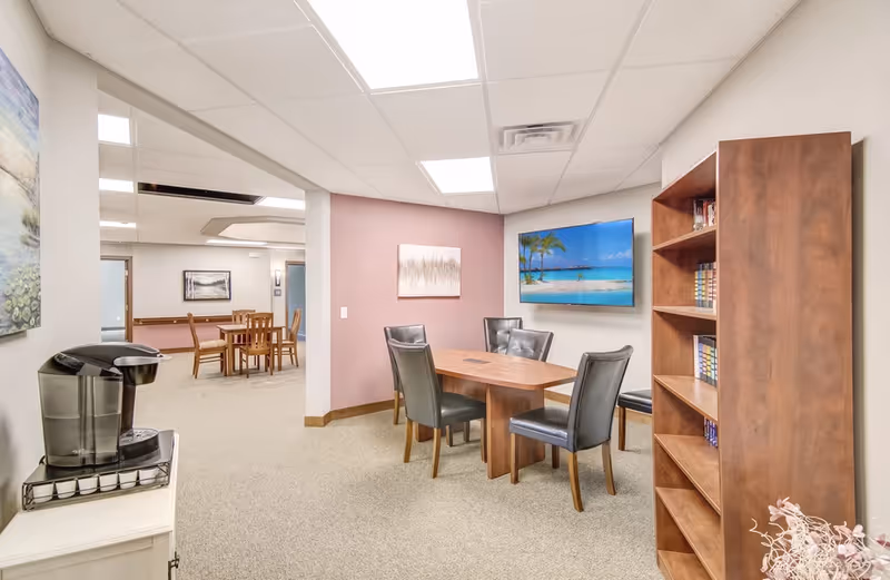 Interior view of a senior living facility common area with a wooden table surrounded by five black leather chairs. A flat-screen TV mounted on a pink accent wall displays a beach scene. To the right, there is a wooden bookshelf with books. In the foreground to the left, there is a coffee maker on a small white cabinet. In the background, another table with chairs is visible in an adjacent room.