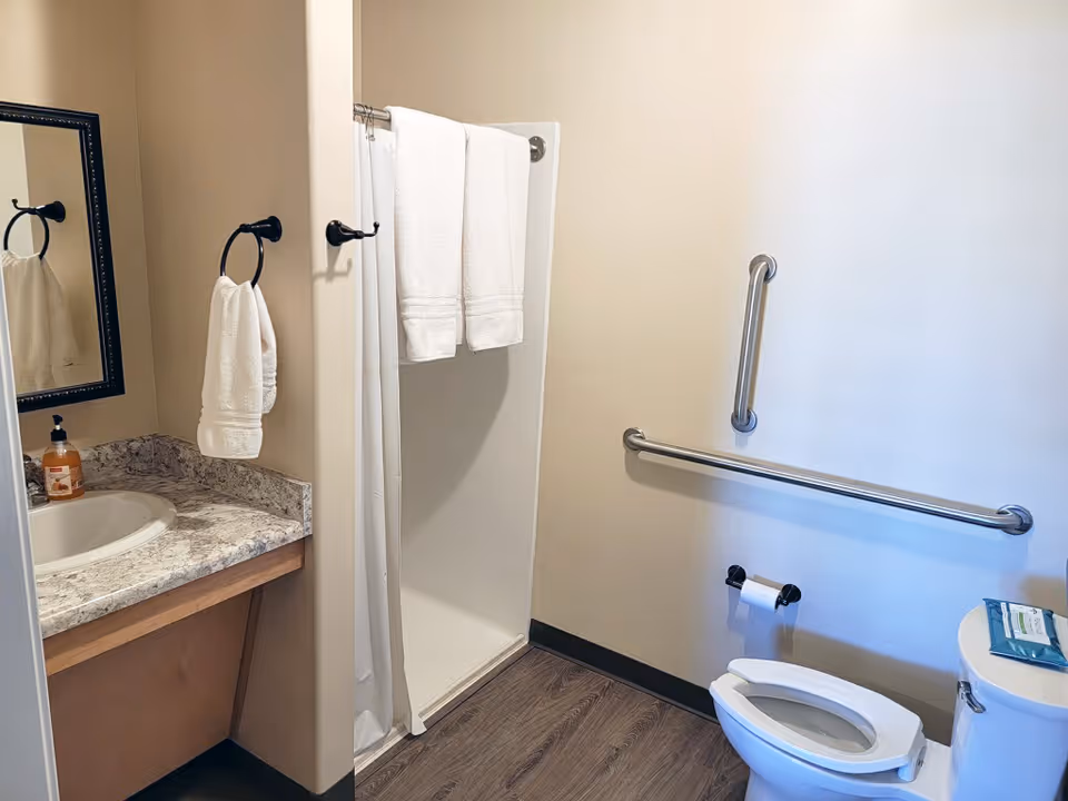 A clean and accessible bathroom featuring a toilet with grab bars on the wall, a shower with a white curtain and two white towels hanging on a rod, a sink with a marble countertop, a mirror, and a hand towel on a ring holder. The floor has wood-like vinyl flooring.