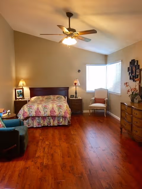 A cozy bedroom with wooden flooring, a bed with a floral bedspread, two nightstands with lamps, a ceiling fan with lights, a cushioned armchair near a window with blinds, and a wooden dresser with decorative items and framed photos on the wall.