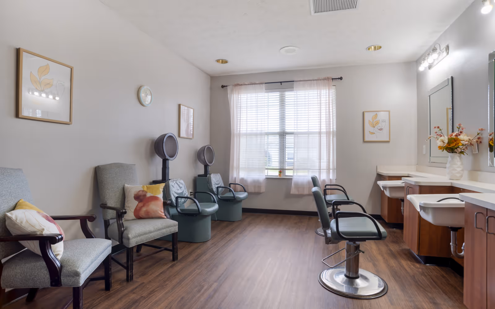 A bright and clean salon area in a senior living facility with two salon chairs in front of mirrors and sinks on the right side, two hair dryer chairs along the left wall, and two cushioned armchairs with decorative pillows. The room has wood flooring, light-colored walls, framed artwork, a clock, and a large window with sheer curtains letting in natural light.