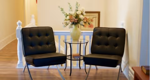 Two black cushioned chairs with metal legs positioned on either side of a small round glass table holding a vase with flowers, set in a hallway with wooden flooring and light-colored walls.