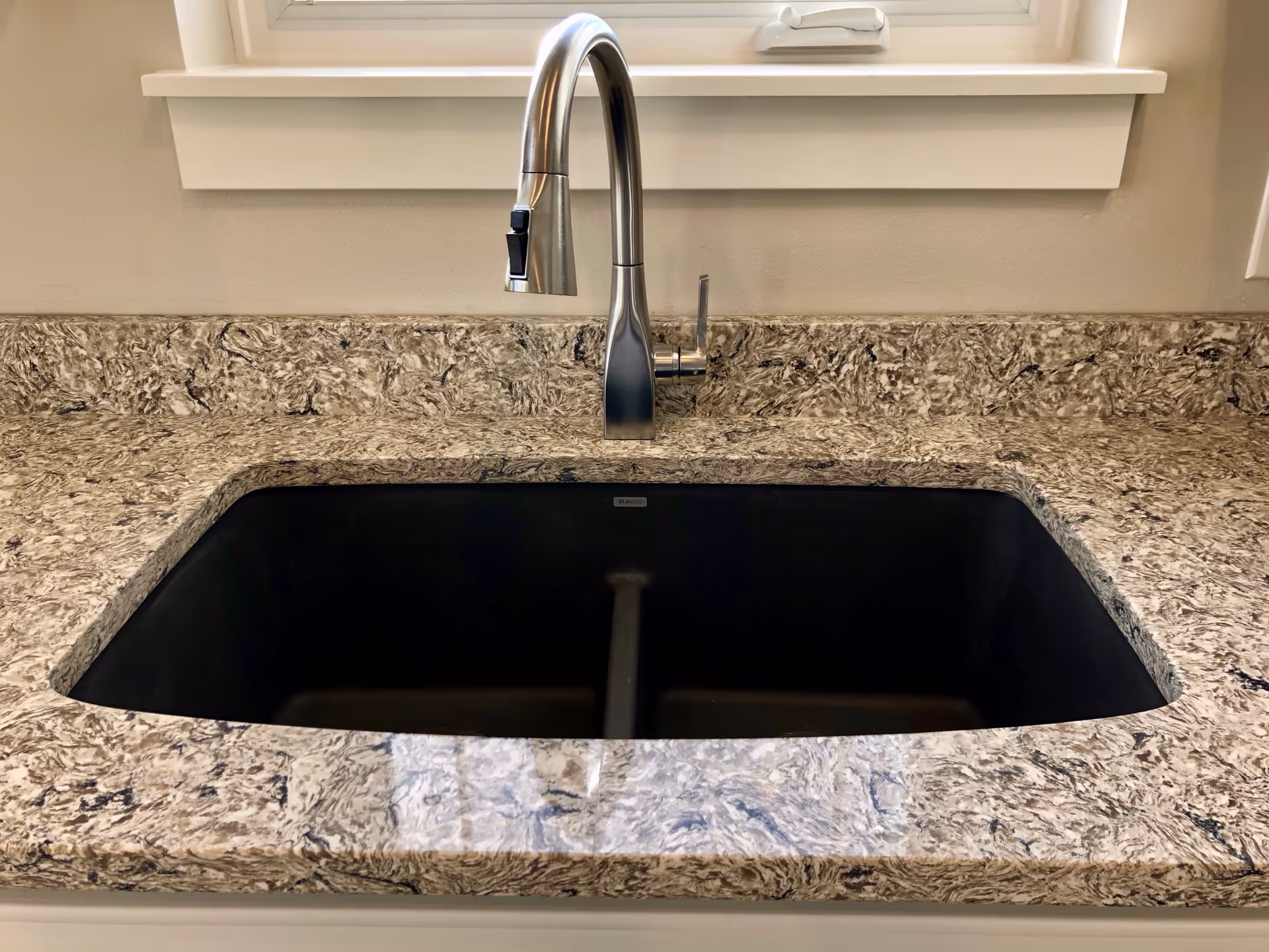 Marble-patterned kitchen countertop with an undermount black sink and a stainless steel faucet beneath a window.