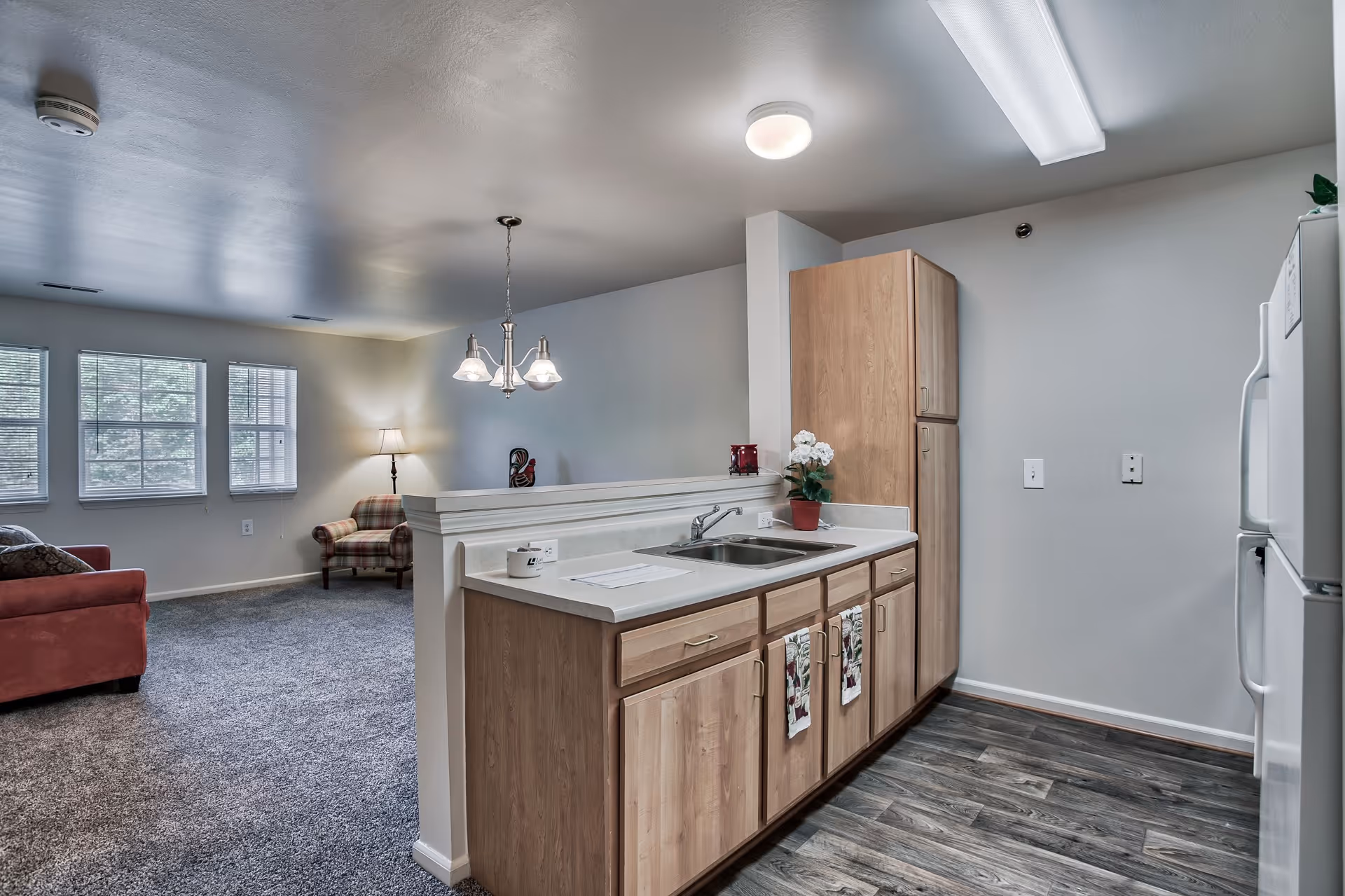 Interior view of a senior living facility showing a kitchen area with wooden cabinets, a double sink, and a white refrigerator. The kitchen has wood-patterned flooring and is adjacent to a carpeted living room area with a red sofa, a plaid armchair, three windows, and a floor lamp. A hanging light fixture is above the kitchen counter.