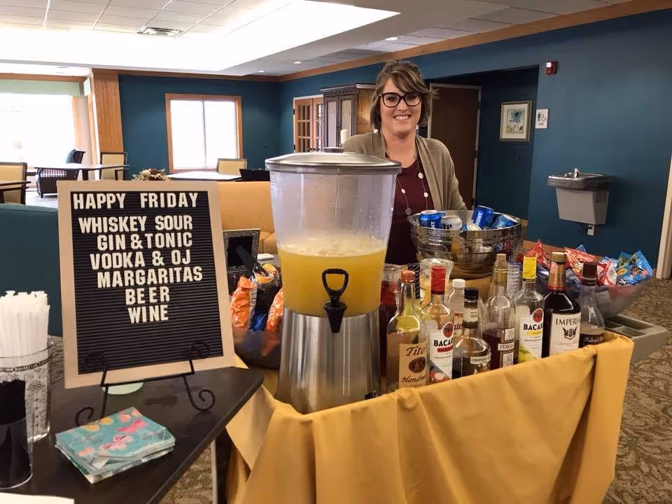 A woman standing behind a table set up with various alcoholic beverages, a large container of yellow drink, snacks, and a sign listing drink options including whiskey sour, gin & tonic, vodka & orange juice, margaritas, beer, and wine in a senior living facility common area.