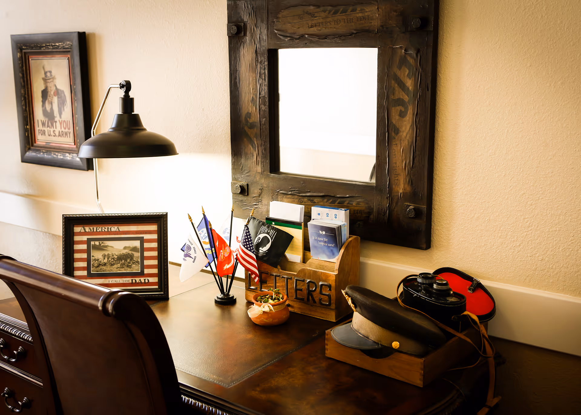 A wooden desk with a vintage military theme, featuring a framed Uncle Sam 'I Want You for U.S. Army' poster, small American and military flags, a wooden letter holder with brochures, a military hat, and a vintage camera. A large rustic mirror hangs on the wall above the desk, and a desk lamp provides warm lighting.