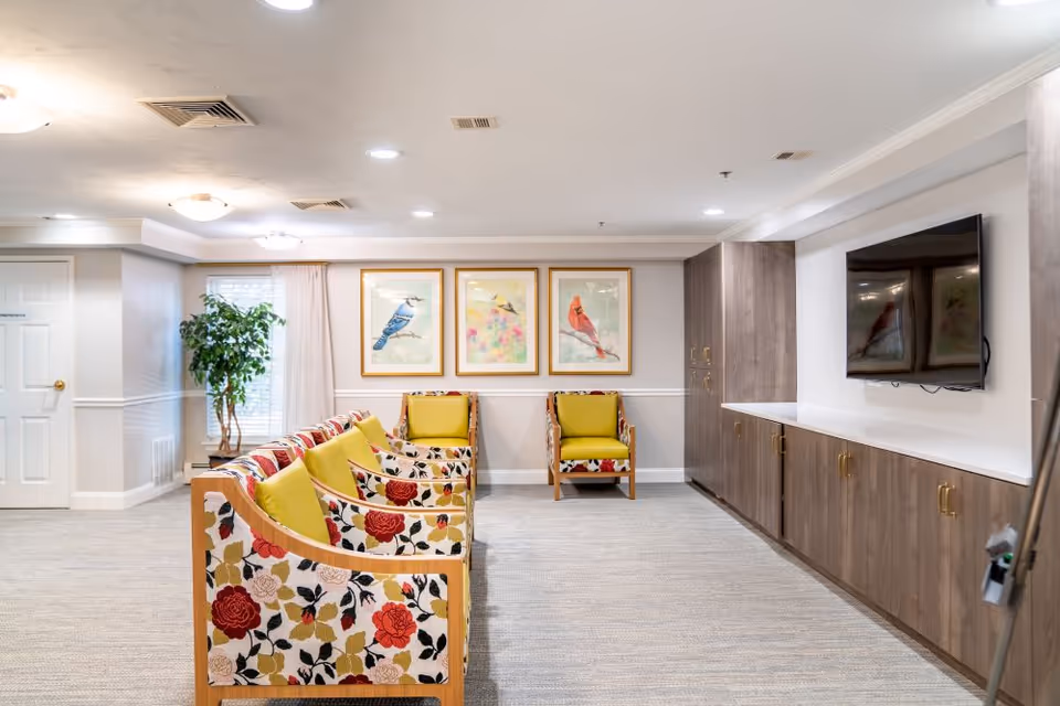A bright and spacious common area in a senior living facility featuring a row of floral-patterned armchairs with yellow cushions facing a wall-mounted flat-screen TV. The room has light-colored walls, a large window with sheer curtains, three framed bird paintings on the wall, a potted plant near the window, and built-in wooden cabinets with gold handles beneath the TV.