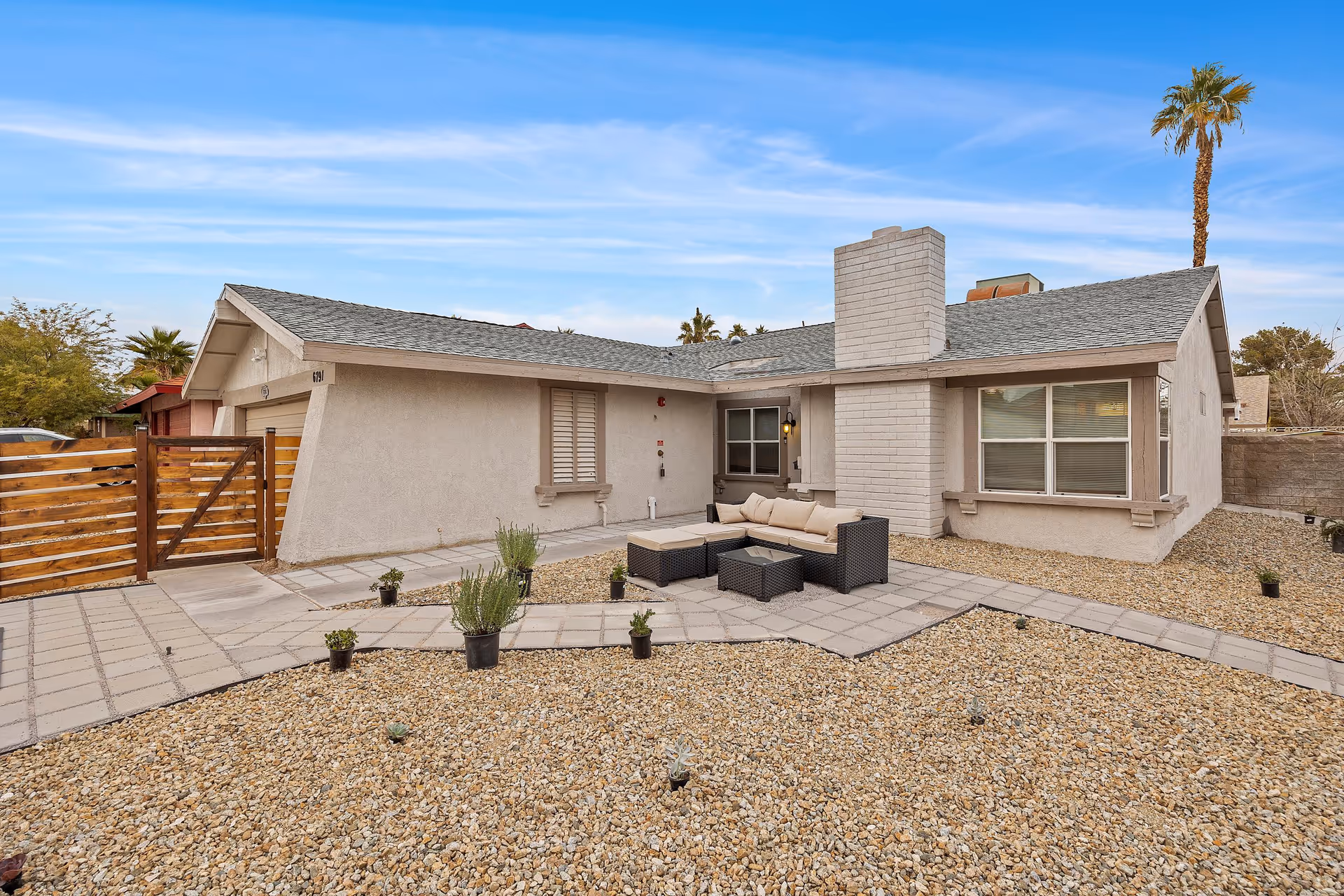 Exterior view of a single-story house with a gray roof and beige walls. The front yard is covered with small rocks and has a paved walkway leading to a wooden gate. There is an outdoor seating area with a cushioned sectional sofa and a glass-top coffee table near a white brick chimney. A tall palm tree is visible in the background under a blue sky with light clouds.