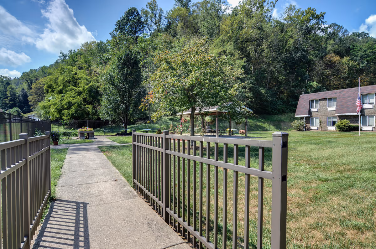 Paved walkway through an open metal gate into a grassy courtyard with a gazebo, trees, and a two-story building with an American flag.