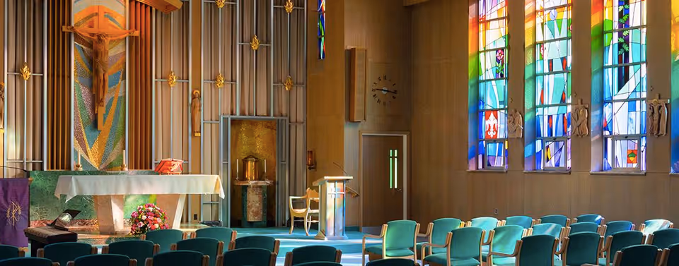 Sunlit chapel interior with an altar, lectern, rows of teal chairs, and stained glass windows.