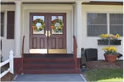 Front entrance of a building with double wooden doors decorated with autumn wreaths. There are steps leading up to the doors with handrails on both sides. To the right of the entrance, there is a window with a flower box containing yellow flowers and a large potted plant below it. The exterior walls are light-colored with dark shutters on the window.