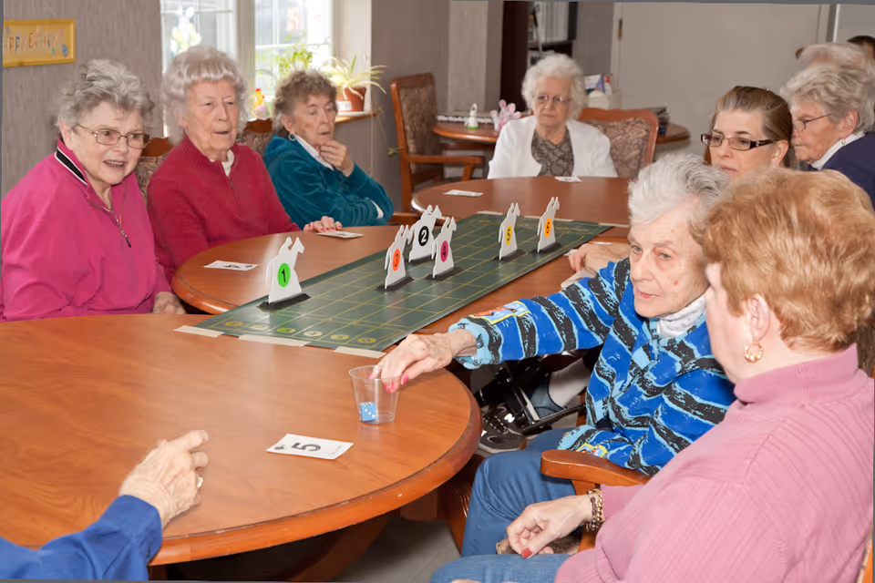 A group of elderly women sitting around a large wooden table playing a board game with numbered horse-shaped markers. One woman is rolling dice into a clear plastic cup while others watch and engage in the activity. The room has windows with natural light and a cozy, communal atmosphere.