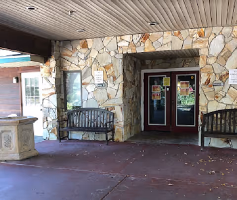 Covered stone-faced building entrance with double glass doors and two wooden benches under a porch.