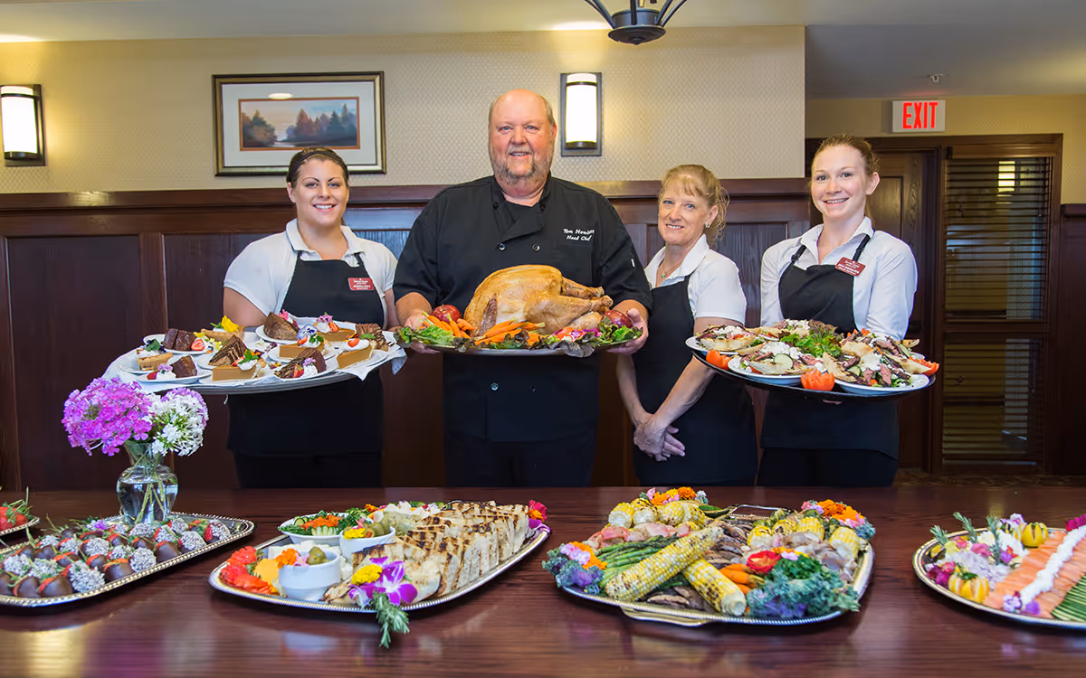 Four catering staff members standing behind a table filled with trays of assorted food including desserts, grilled items, vegetables, and a large roasted turkey. The staff are smiling and wearing black aprons, with the chef in the center holding the turkey. The setting appears to be an indoor dining or serving area with wood paneling and a framed picture on the wall.