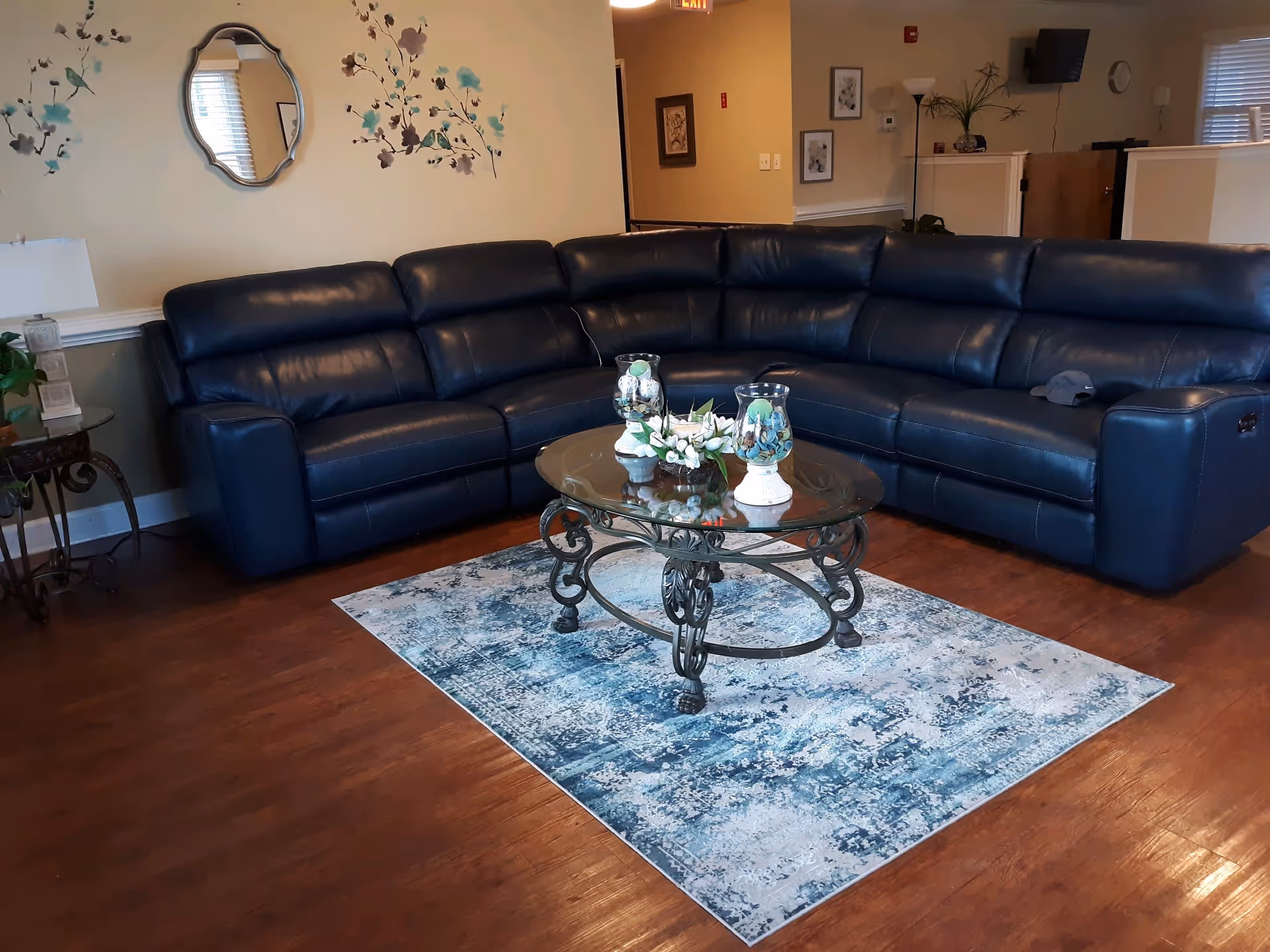 A living room with a large dark blue leather sectional sofa arranged around a glass coffee table with ornate metal legs. The coffee table holds decorative items including two glass candle holders and a floral arrangement. The floor is wooden with a blue and white patterned rug under the coffee table. The walls are light-colored with floral wall decals and a decorative mirror. There is a side table with a lamp and a plant, and in the background, there are framed pictures, a floor lamp, and a wall-mounted TV.