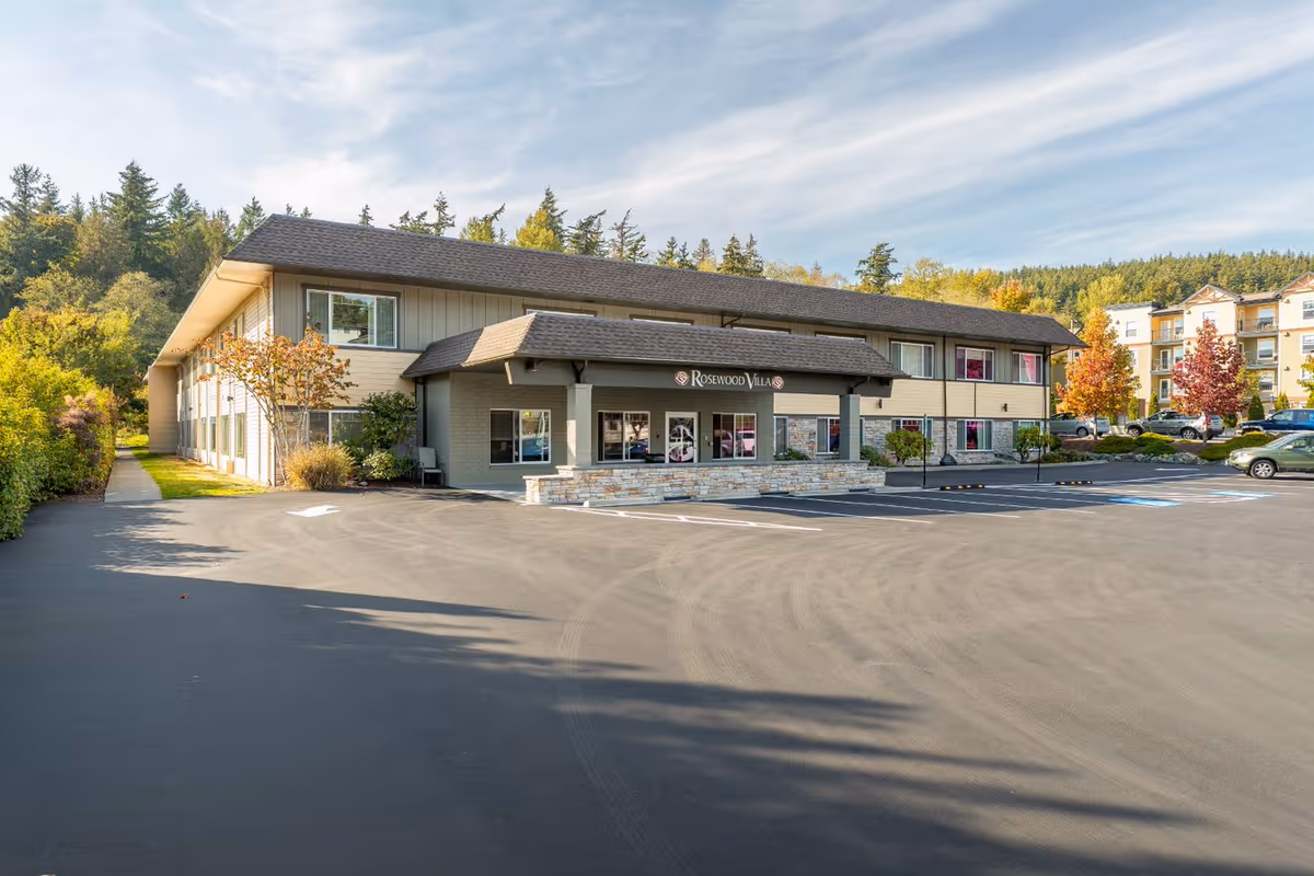 Exterior view of Rosewood Villa, a two-story senior living facility with a large parking lot in front, surrounded by trees and greenery under a partly cloudy sky.