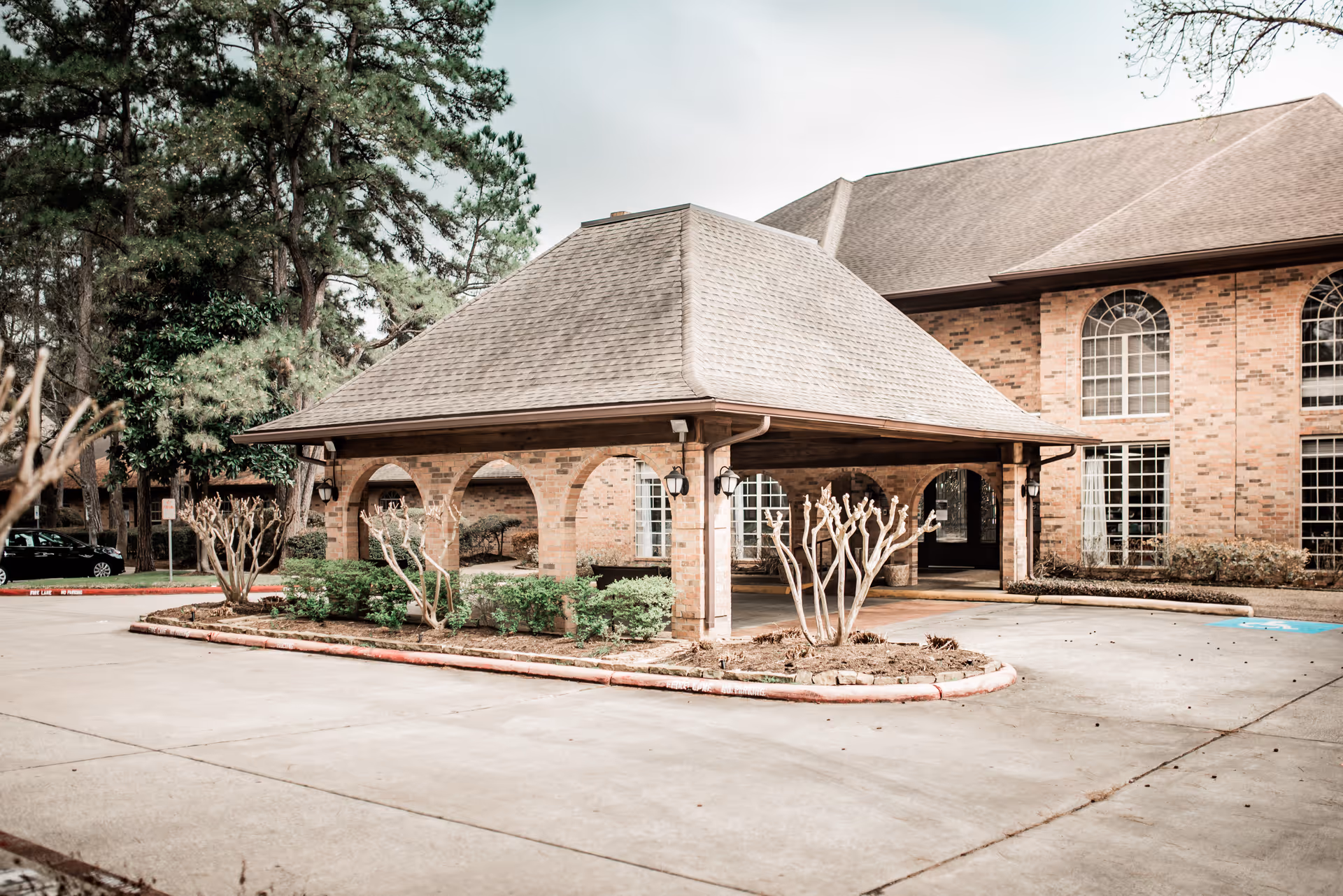 Exterior view of a brick building with a large covered entrance supported by brick arches, surrounded by trimmed bushes and trees, under an overcast sky.