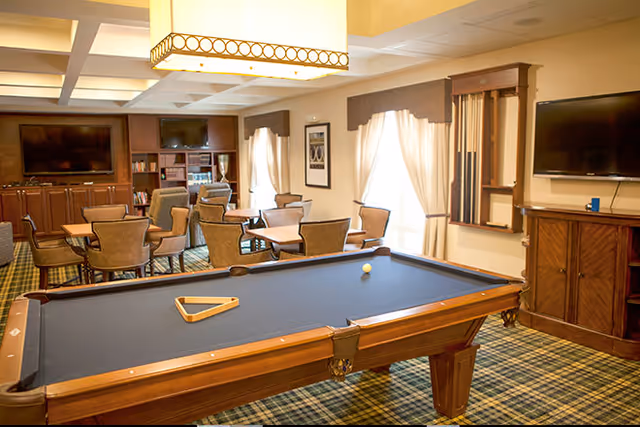 A recreational room with a pool table in the foreground, a rack with pool cues on the wall, multiple tables and chairs arranged for seating, two large flat-screen TVs mounted on the walls, and a cabinet with shelves and decorative items. The room has large windows with curtains and a patterned carpet floor.