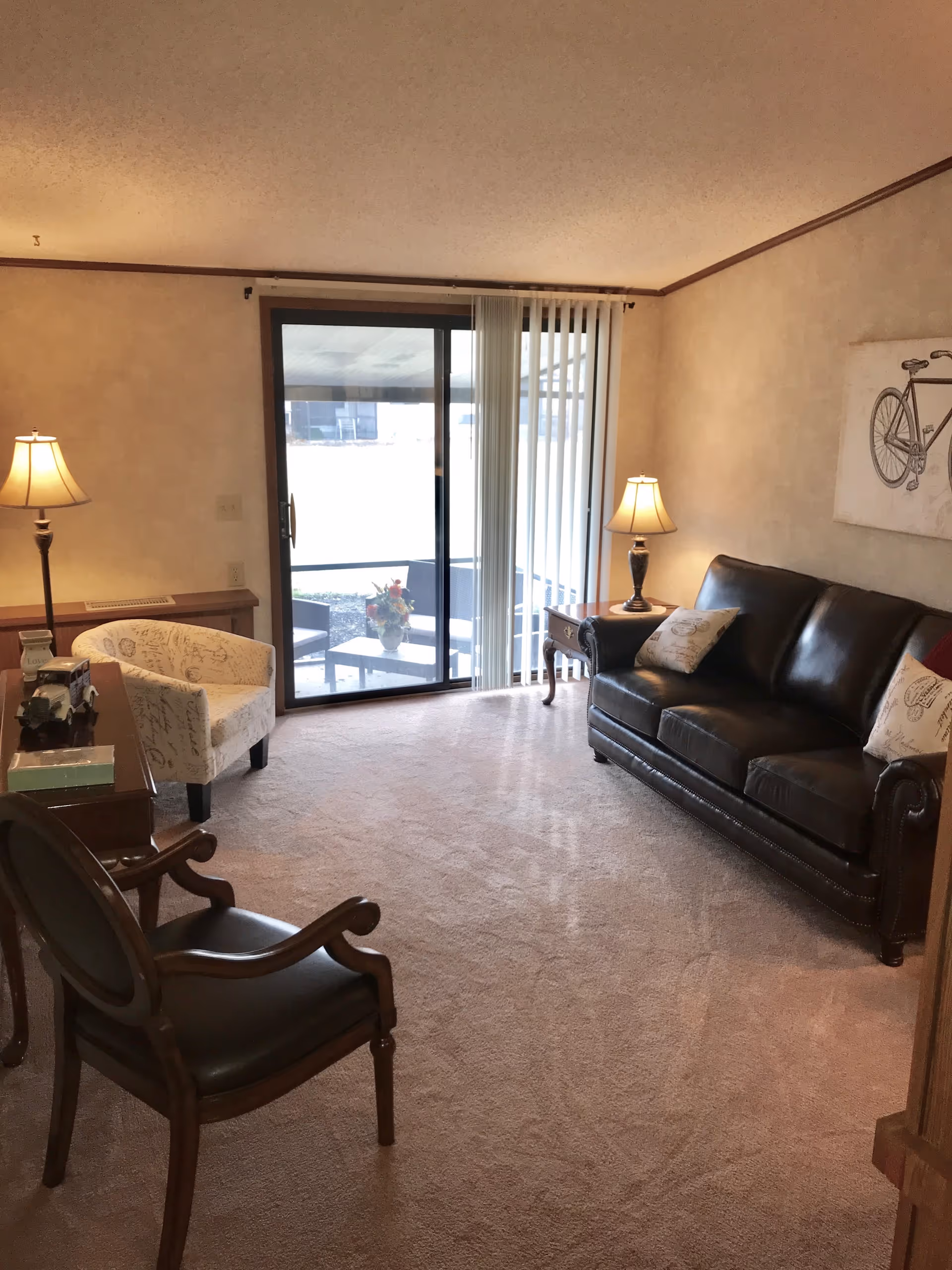 A cozy living room with a dark leather sofa adorned with two decorative pillows, a wooden side table with a lamp, a patterned armchair, and two wooden chairs with dark cushions. There is a sliding glass door with vertical blinds leading to an outdoor patio area with chairs and a table with a flower arrangement. The room has beige carpet and walls with a bicycle artwork hanging on the wall.