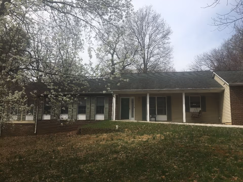 Front exterior view of a single-story assisted living facility building with a covered porch, green door, several windows with shutters, a porch swing, and trees with white blossoms in the foreground.