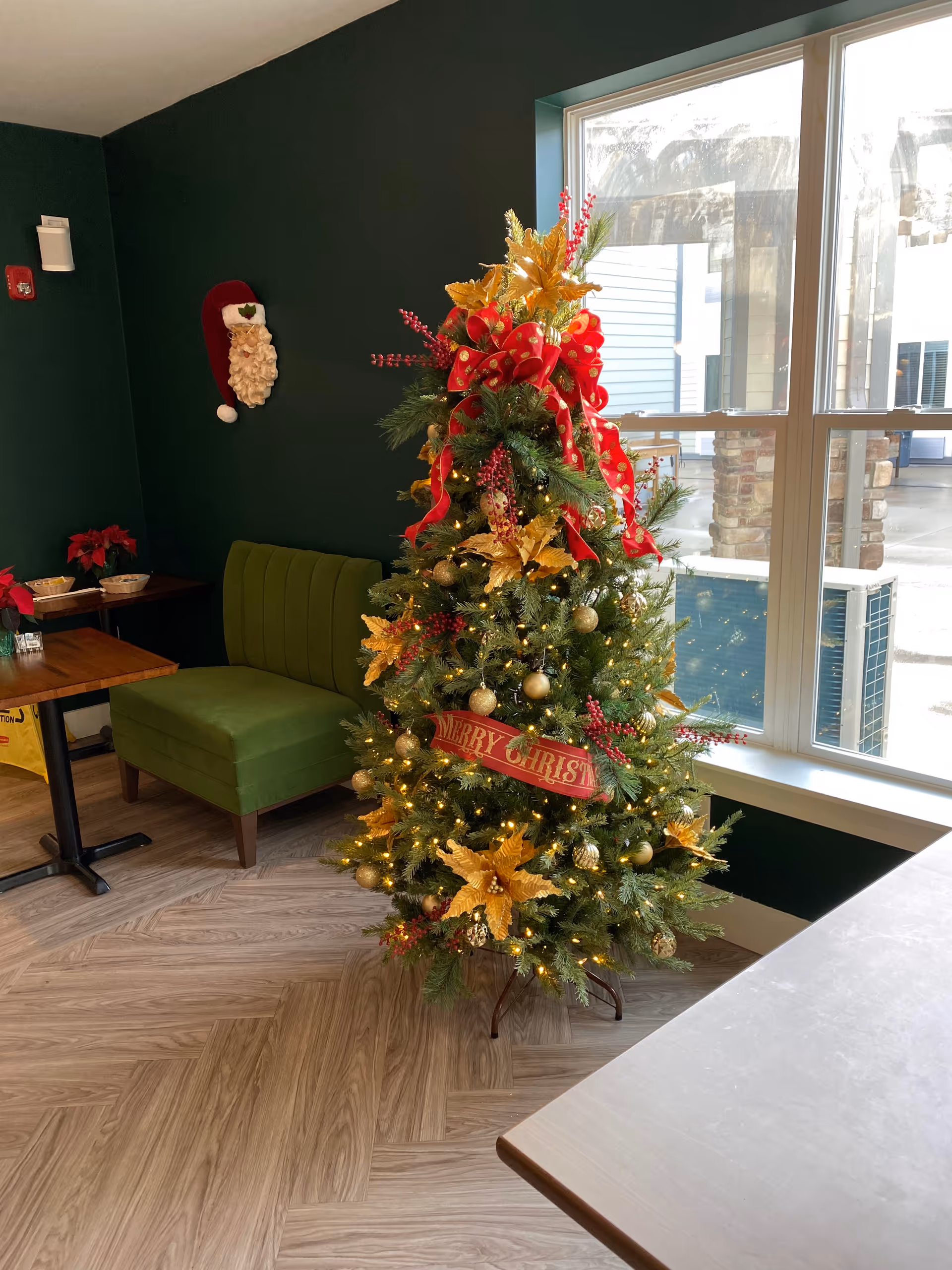 Christmas tree decorated with gold poinsettias and a 'Merry Christmas' sign in a communal dining area with green seating by a large window.