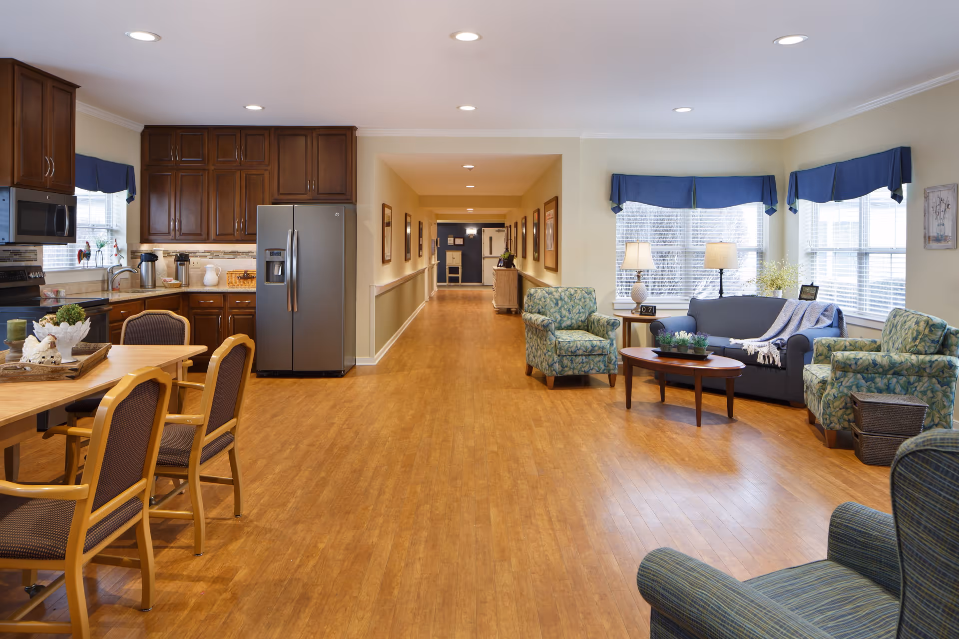 A spacious and well-lit interior room featuring a kitchen area with wooden cabinets, stainless steel refrigerator, microwave, and a dining table with chairs on the left. On the right side, there is a cozy living area with a sofa, two armchairs, a coffee table, and lamps near large windows with blue valances. A long hallway extends from the center of the room, decorated with framed pictures on the walls.