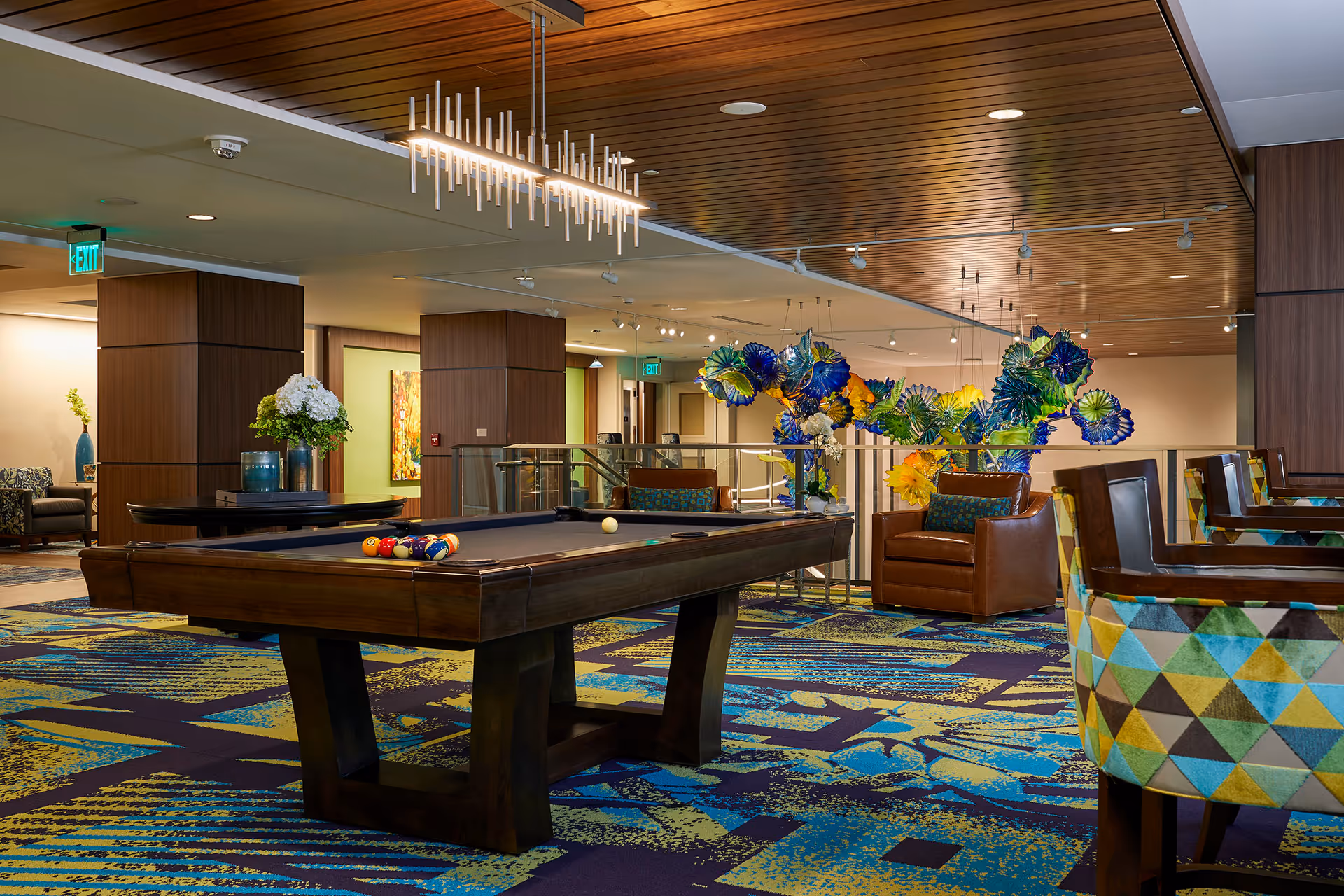 A stylish senior living common area featuring a pool table with balls set up, colorful patterned carpet, modern armchairs with geometric upholstery, a brown leather chair, and a decorative glass sculpture in blue, green, and yellow hues. The ceiling has wooden paneling with contemporary lighting fixtures, and the space is well-lit with recessed lights and exit signs visible in the background.