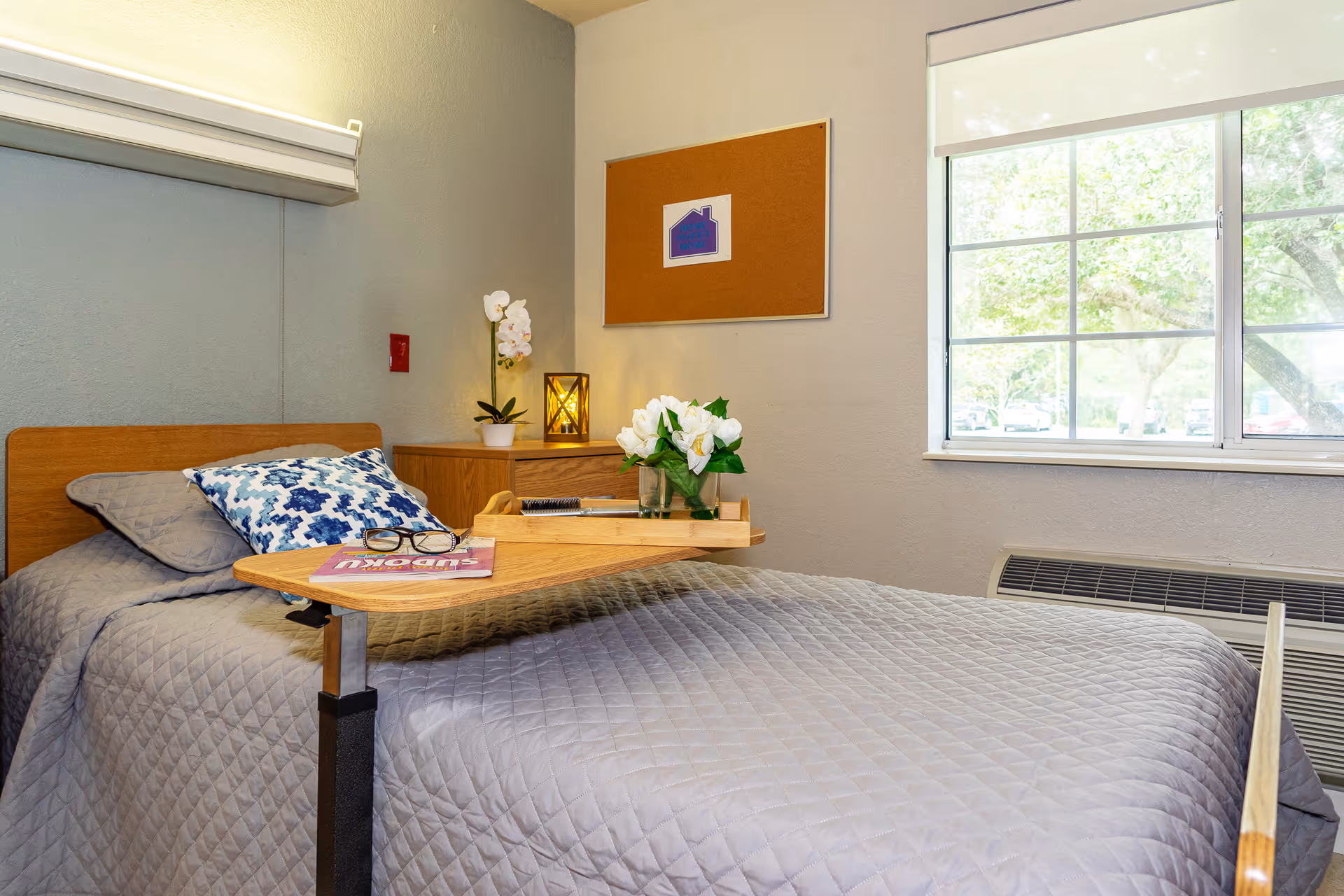 A neatly made single bed with a gray quilted bedspread and a blue patterned pillow in a nursing home room. A wooden overbed table holds a magazine, eyeglasses, a notebook, and a vase with white flowers. There is a wooden nightstand with a potted orchid and a decorative lantern. A corkboard with a small house-shaped decoration hangs on the wall. A large window with a white blind lets in natural light, showing trees outside.