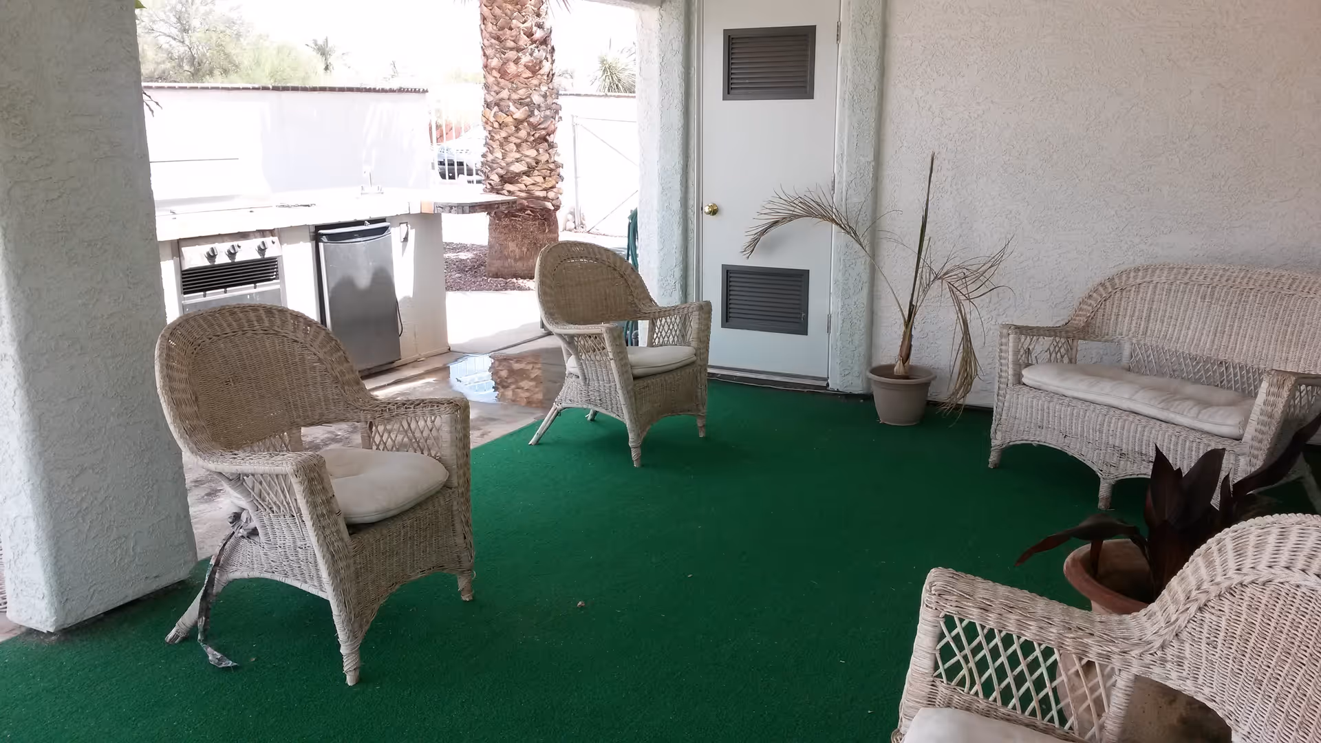 Covered outdoor patio area with green carpet flooring, featuring white wicker chairs and a loveseat with cushions. There are potted plants and a built-in outdoor kitchen with a grill and mini fridge visible in the background.