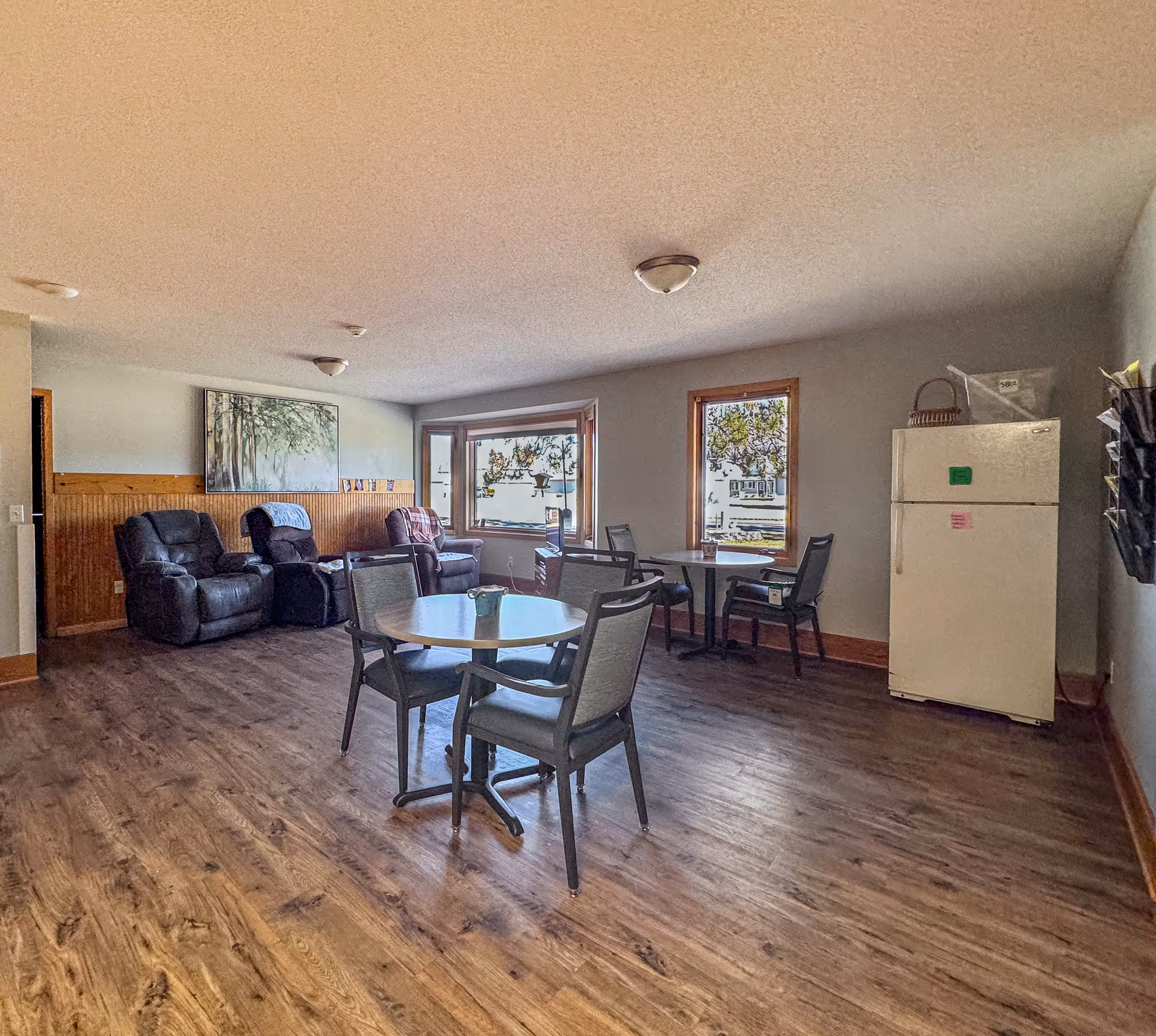 A common area in a senior living facility with wood flooring, two round tables each surrounded by four chairs, three recliner chairs against the wall, a large window with a view outside, and a white refrigerator in the corner.