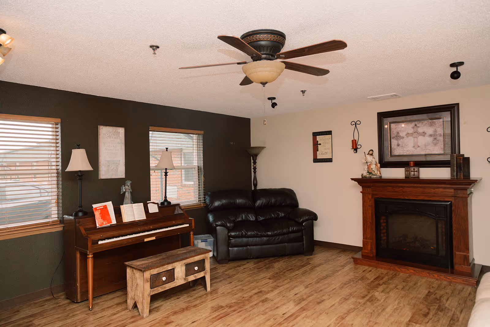 Cozy living room with a piano and bench, leather loveseat, wooden fireplace mantel, ceiling fan, and lamps.