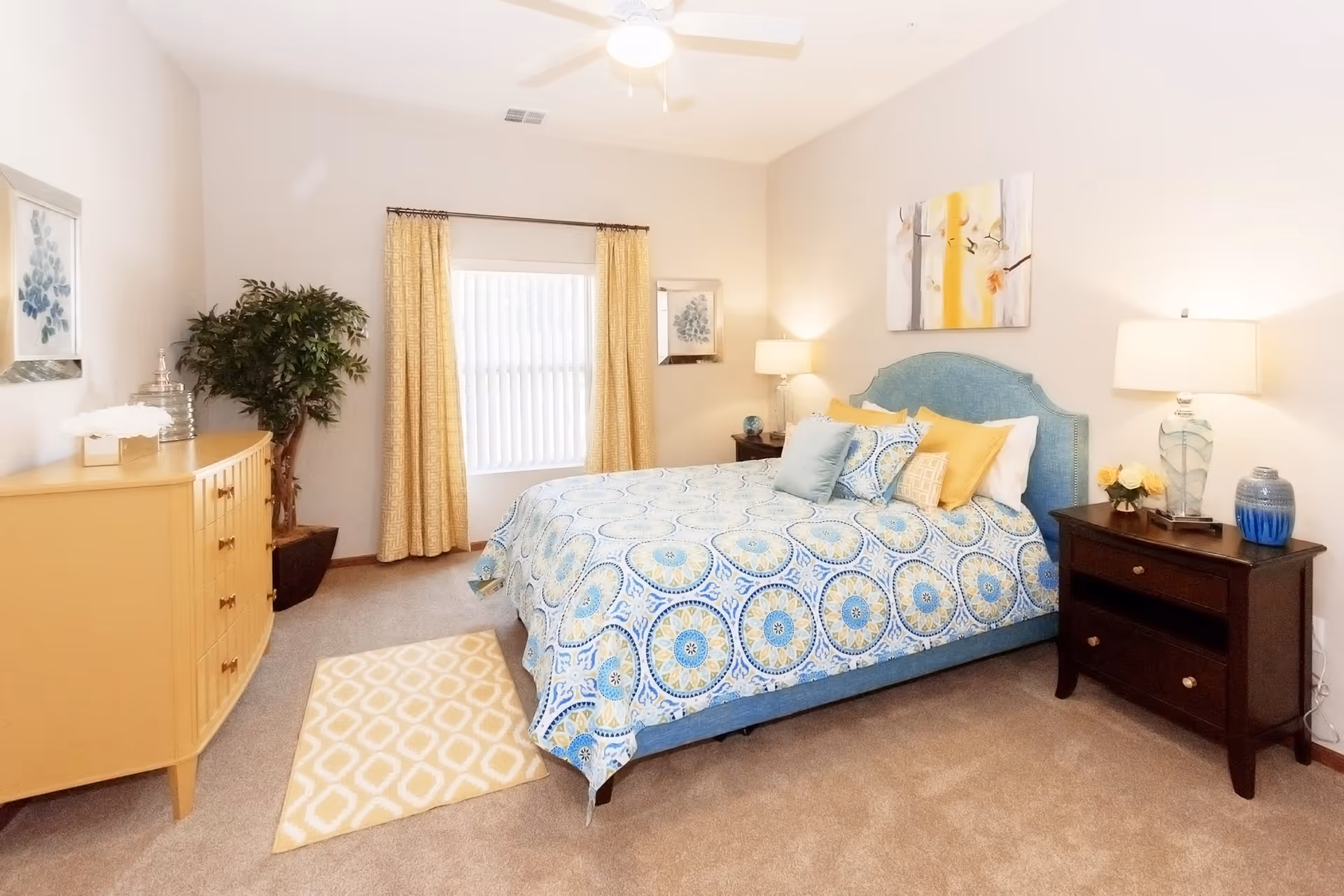 A bright and cozy bedroom featuring a bed with a blue and yellow patterned bedspread and multiple pillows. The room has beige carpet, a ceiling fan with light, two dark wooden nightstands with lamps and decorative items, a yellow dresser, a potted plant, yellow patterned curtains, and framed artwork on the walls.
