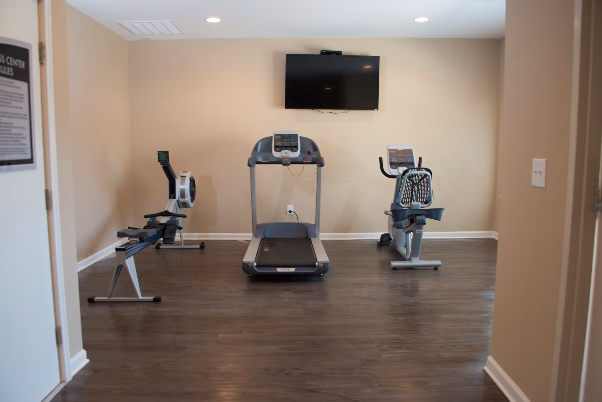 A small fitness room with three exercise machines: a rowing machine on the left, a treadmill in the center, and a recumbent bike on the right. A flat-screen TV is mounted on the beige wall above the treadmill. The room has wood flooring and recessed ceiling lights.