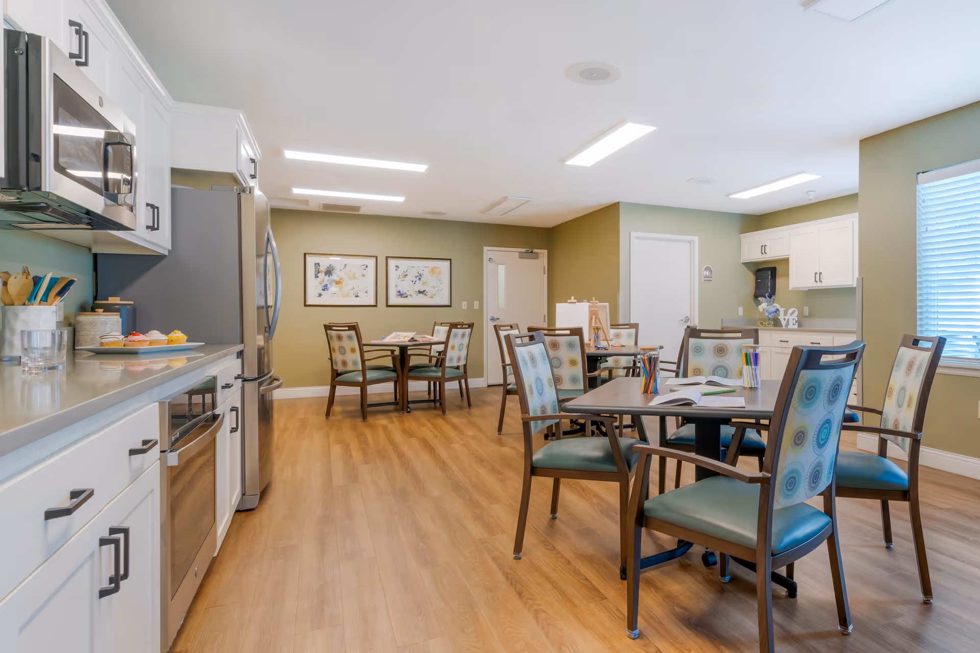 A bright and clean communal dining and kitchen area with wooden flooring, white cabinets, stainless steel appliances, and several tables with chairs featuring patterned upholstery. The walls are painted green and decorated with framed artwork, and a window with blinds allows natural light into the room.
